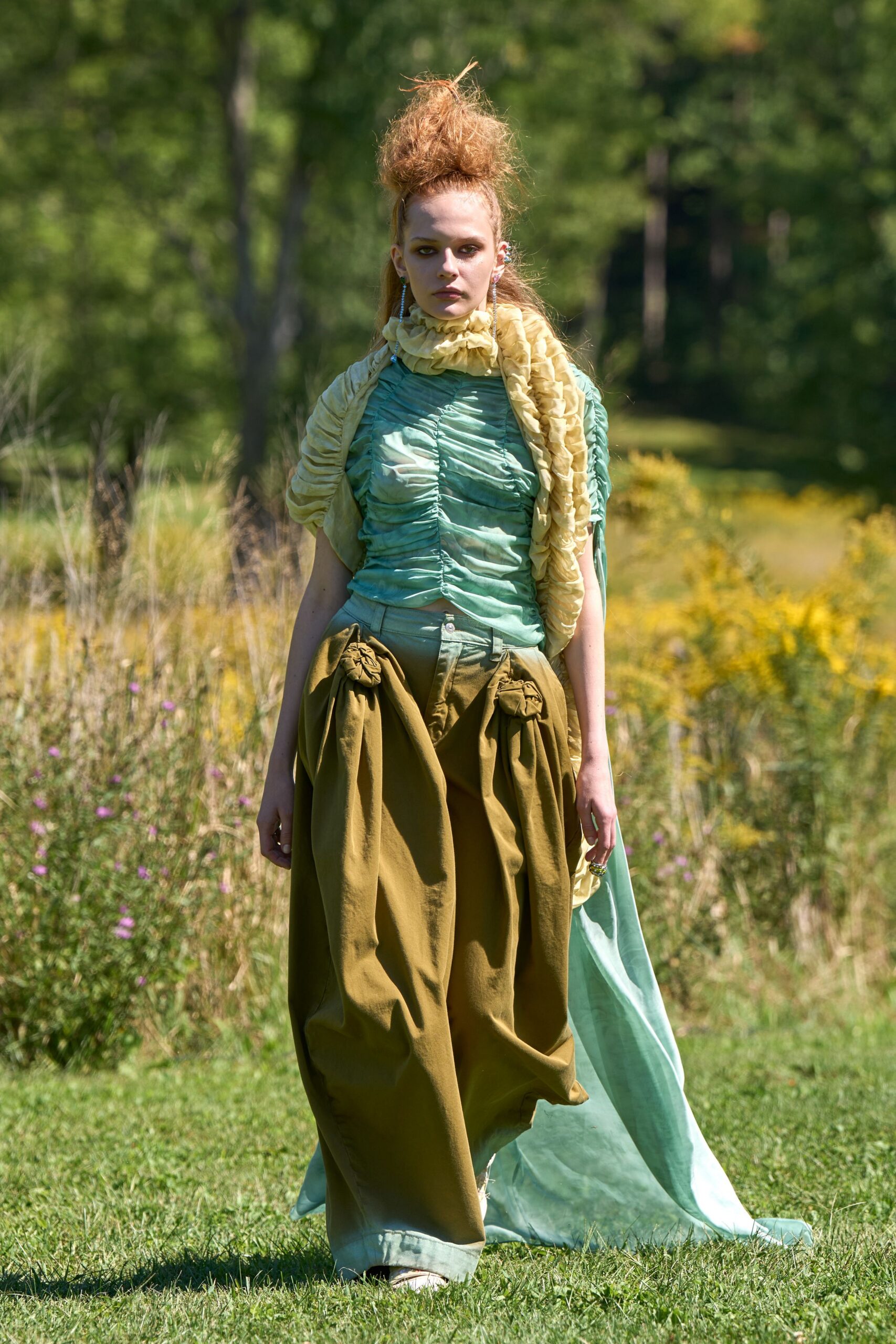 Butter-yellow organza scarf, bubbled into soft ruffles around the neck. Seafoam ruched top with a puckered center seam. Olive balloon trousers, hip swags knotted into rosettes. Pale aqua panel dragging loose behind the hem. Long crystal drops. Crossing a meadow after the garden wedding has thinned out.