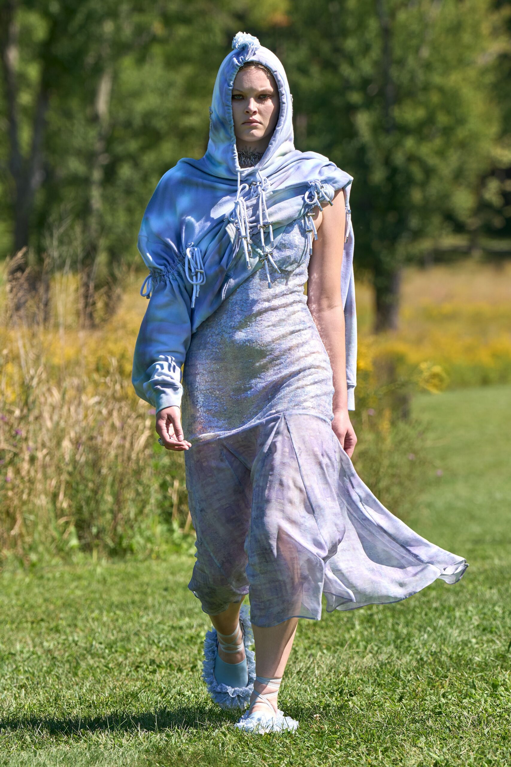 Powder-blue hoodie draped askew, threaded with dangling white drawcord loops. Iridescent slip dress with a crushed surface and sheer fishtail hem. Dark spiked choker tight at the throat. Pale wrap shoes with frilled edges brushing the grass. Crossing a meadow after rain, when the hem keeps gathering water.