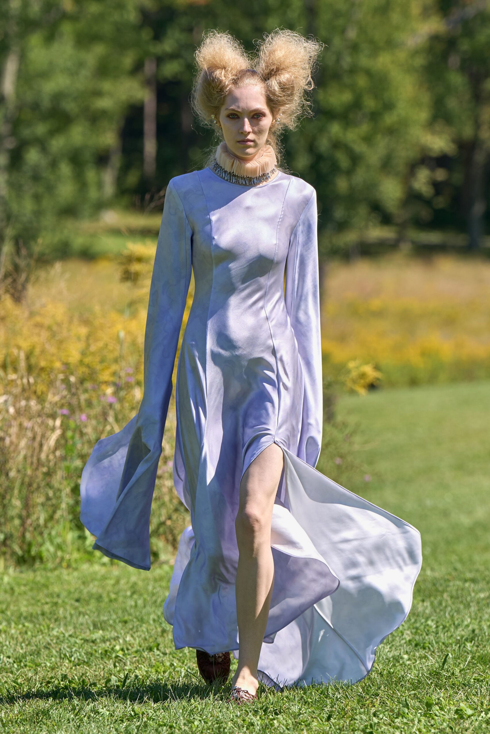 Pale lilac satin dress with fluted sleeves and a wind-lifted thigh slit. Peach pleated organza ruff, ringed with dangling silver spikes. Pointed flats in brown snakeskin. Crossing a meadow after the garden ceremony has already turned strange.