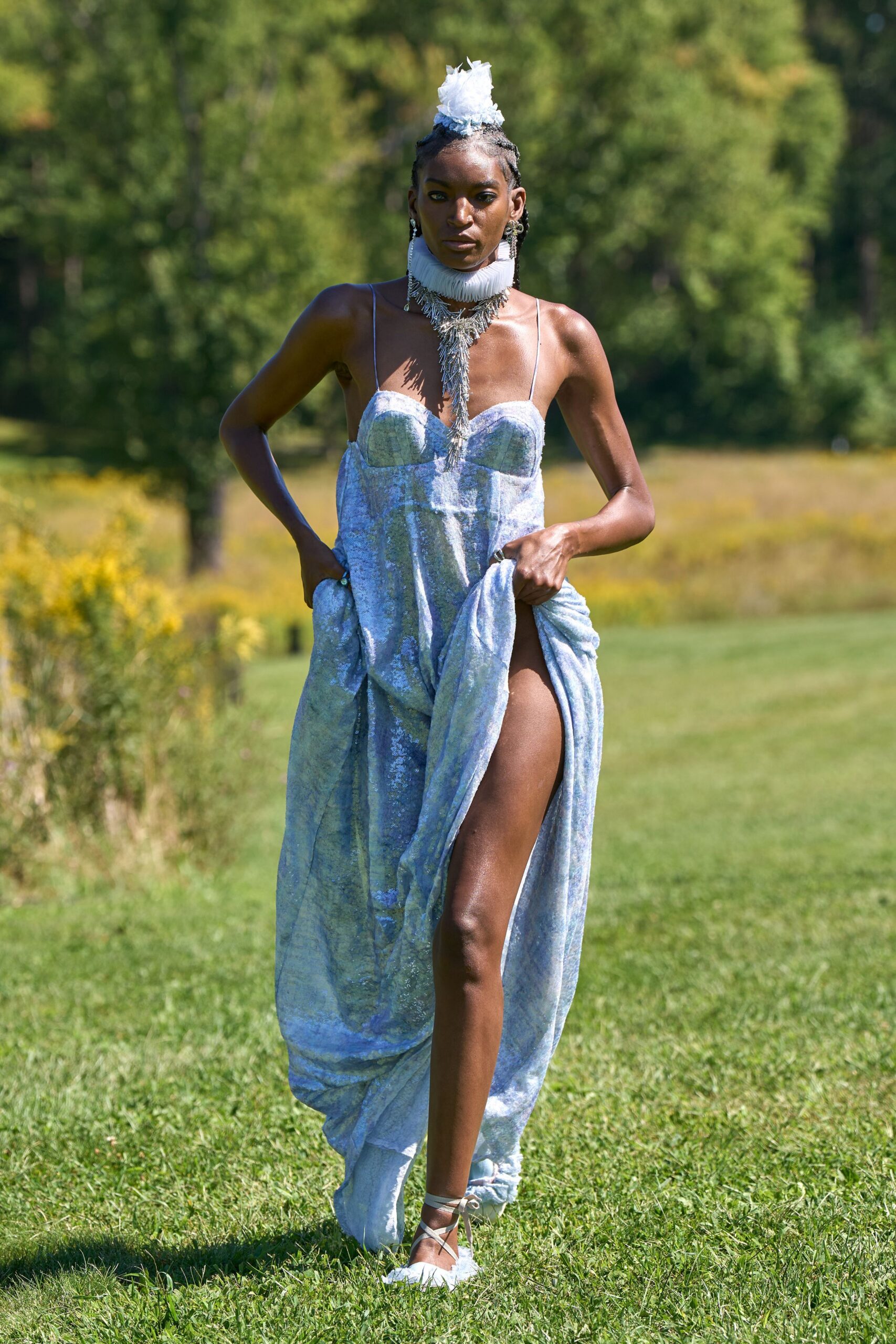 Crushed white tulle bloom pinned over tight cornrows. Pleated white ruff with a sharp cascade of silver fringe. Ice-blue sequin gown with molded cups and a gathered thigh-high opening. White lace-up flats with feathery tufts at the toe. Crossing a wet lawn after the garden vows, shoes sinking first.