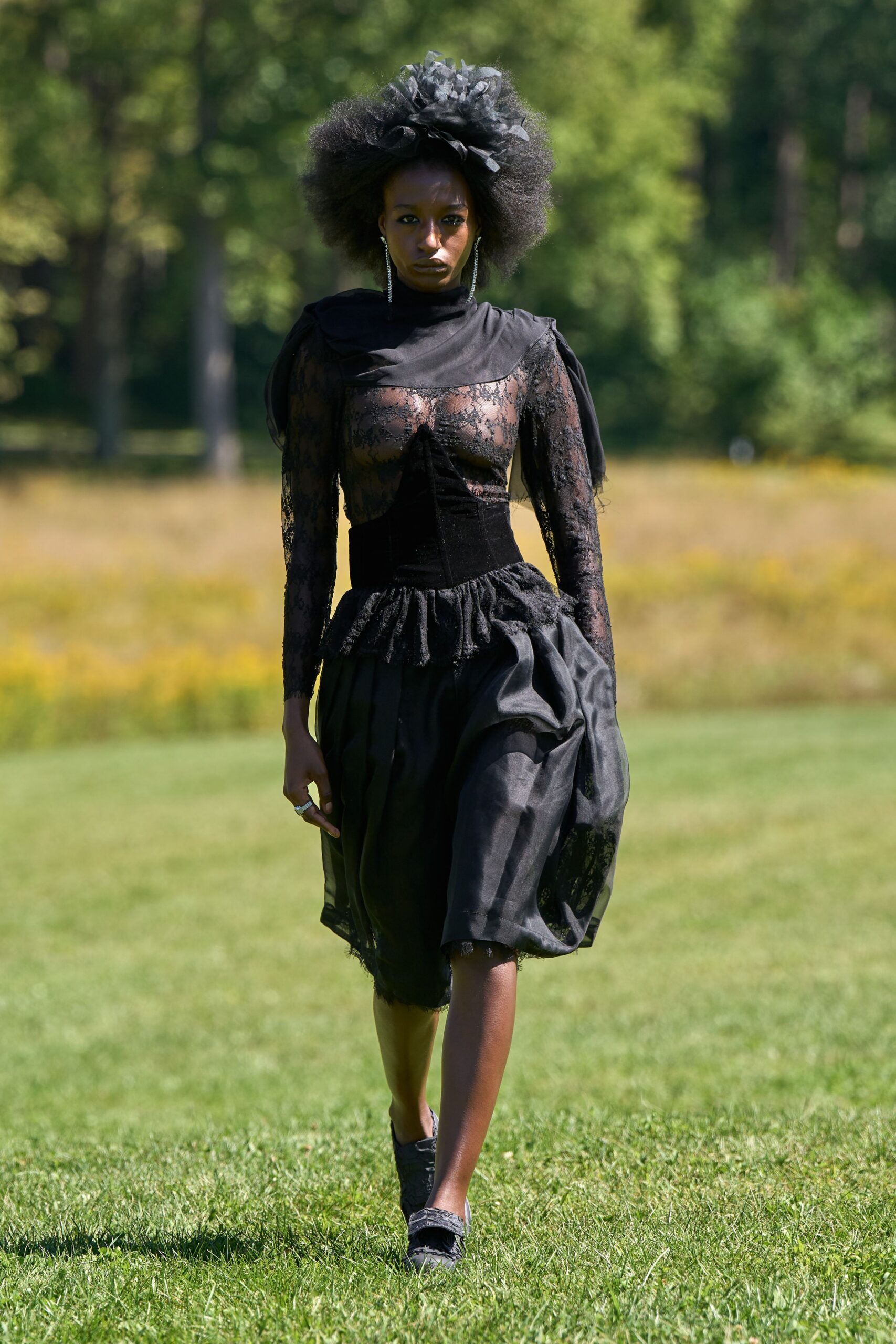 Black organza rosette perched over a halo of natural hair. Sheer black lace bodice over nude lining, velvet corset waist and puffed overskirt. Long rhinestone strands brushing the jaw. Black moire flats with a frayed edge. Crossing the lawn after a memorial service, sun too bright for mourning.