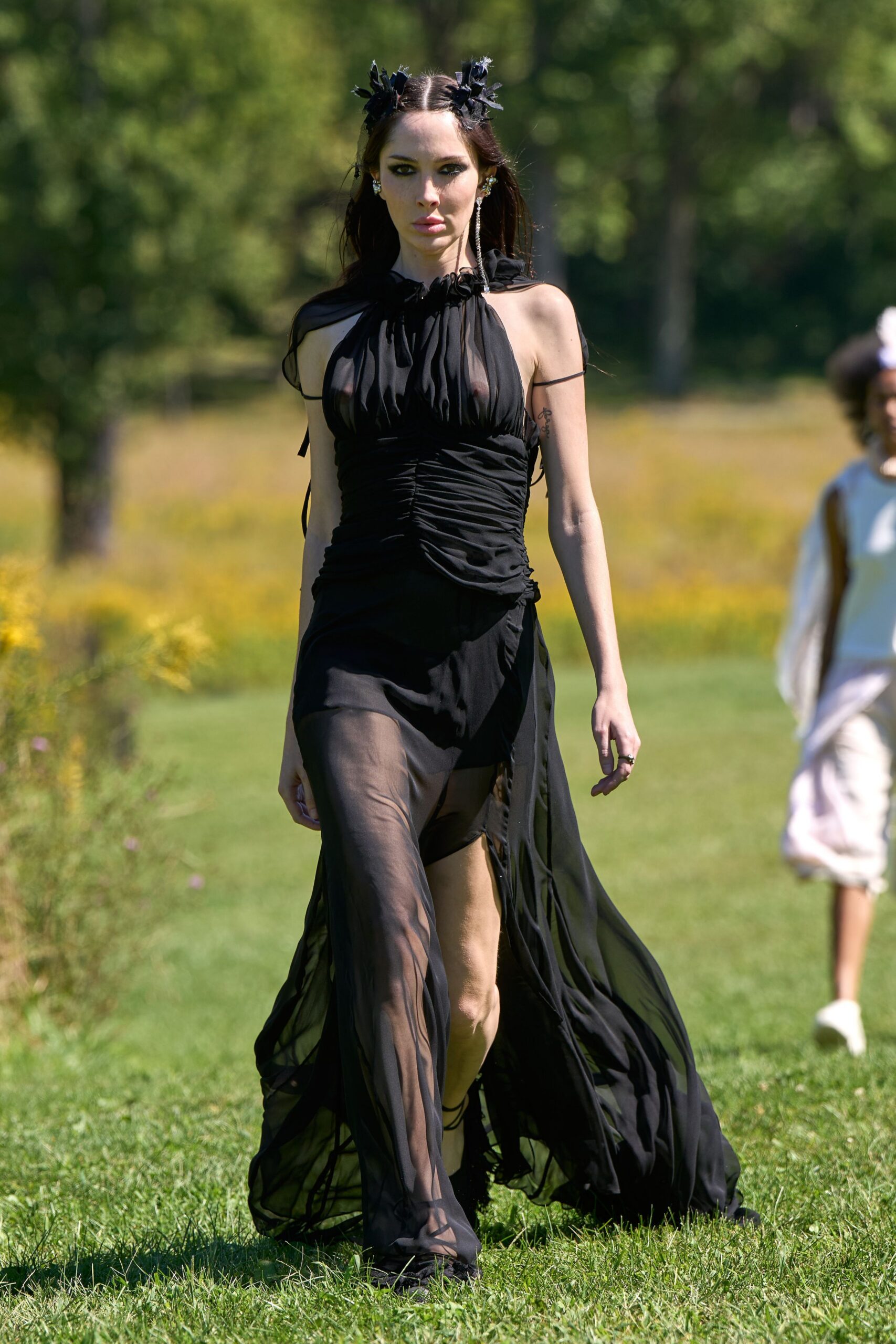 Two black petal clusters pinned beside the center part. Black chiffon halter dress, ruched through the torso with a sheer split skirt. Built-in black shorts showing beneath the gauzy front panels. Black lace-up sandals winding over the calves. Crossing a meadow after the wedding guests have drifted back inside.