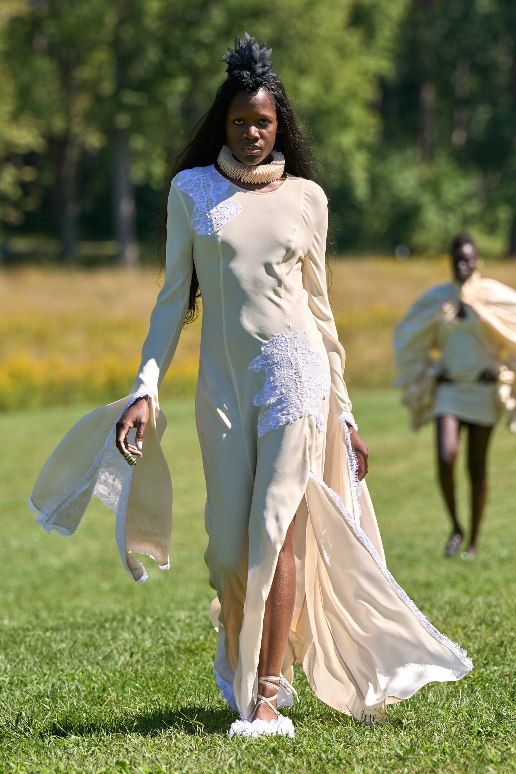 Black tulle flower pinned upright at the crown. Concertina-folded cream collar circling the neck like paper fan blades. Buttercream column gown with white lace appliques and frayed lace edging. Cream lace-up sandals with fluffy white pommed toes. Crossing a manor lawn before the vows, grass still wet underneath.