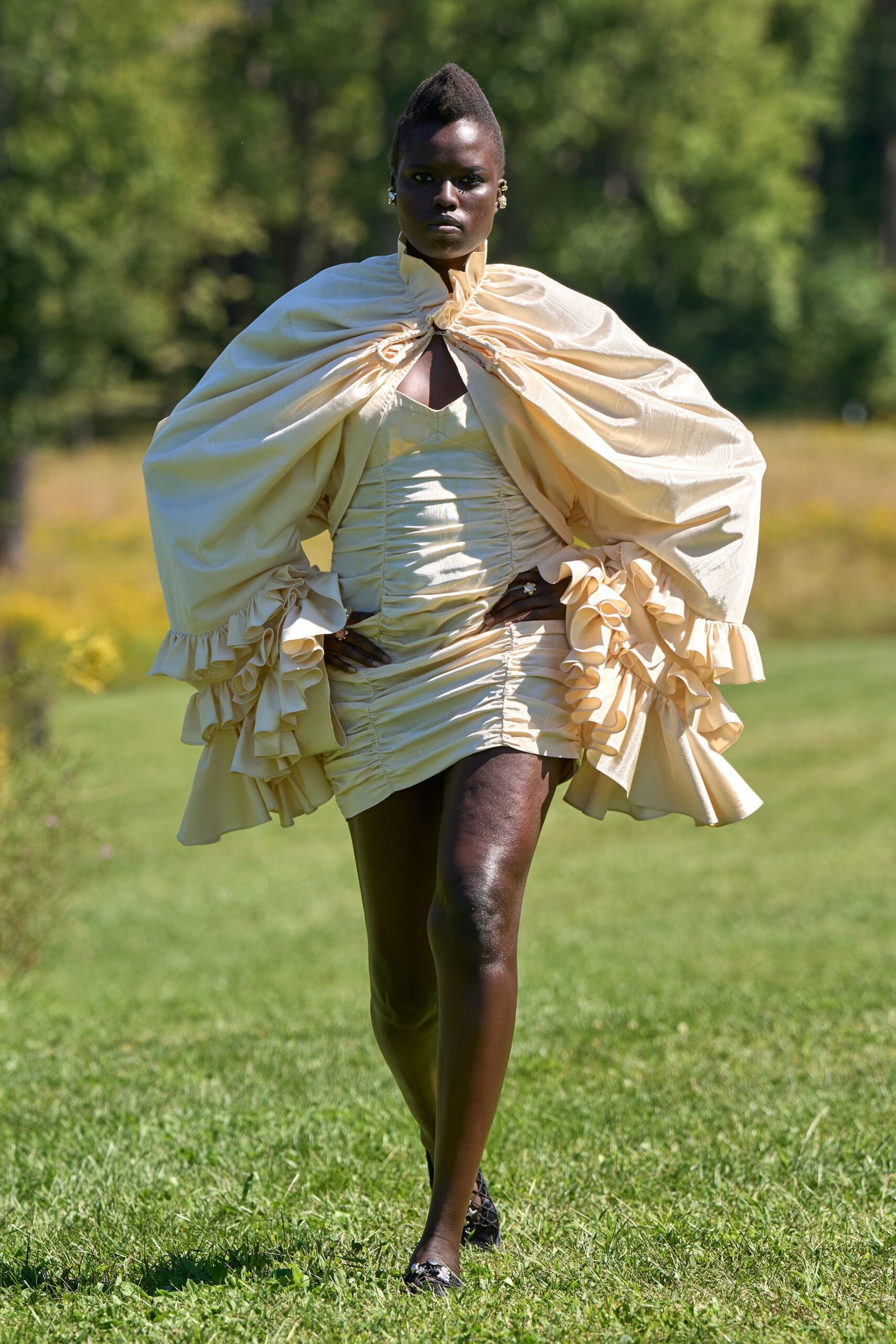 Buttercream taffeta cape, drawn tight at the throat with looping cords. Matching ruched mini dress with a peaked bust and puckered center seam. Black lattice flats. Crossing a sunburnt lawn toward a tented wedding after the champagne started.