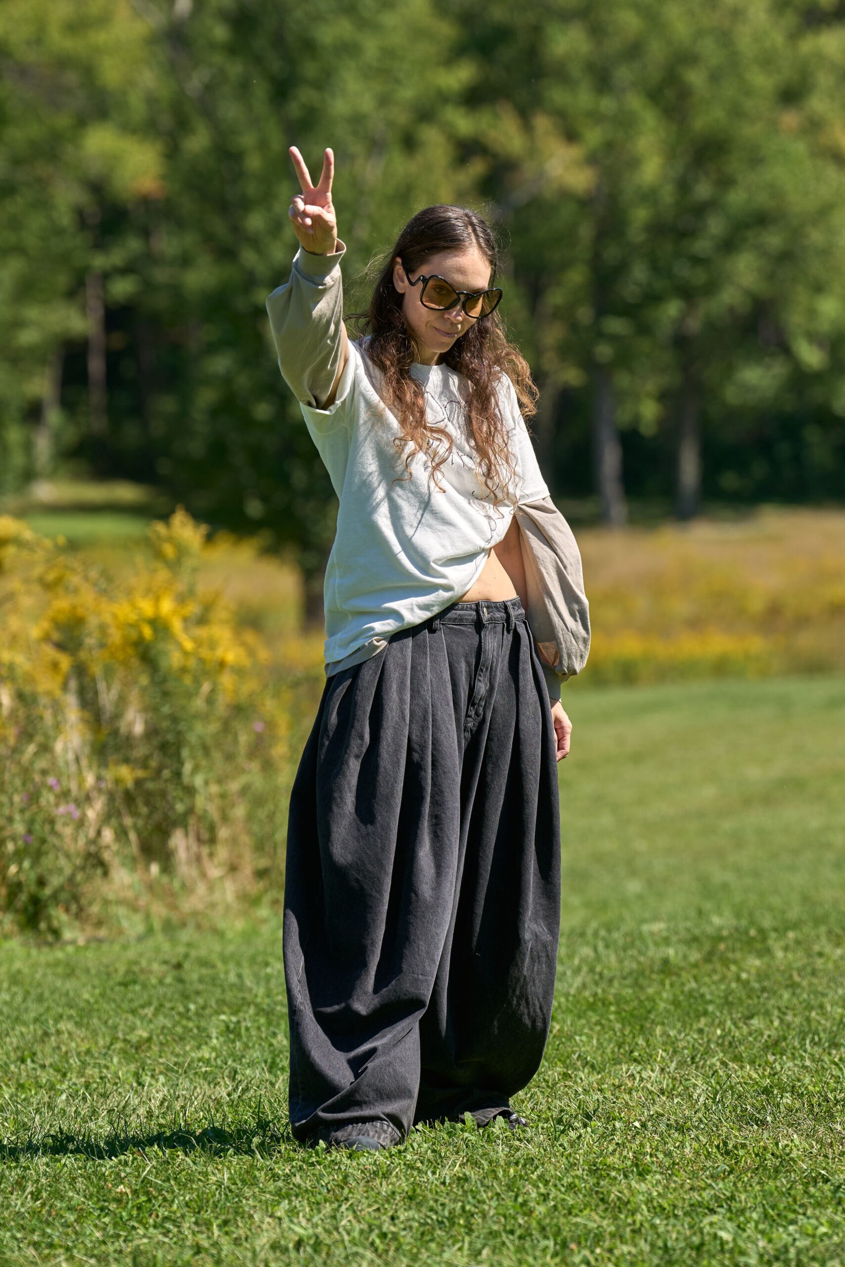 Amber shield sunglasses with thick black curved rims. Stone jersey sleeves extending past an oversized white crewneck tee. Charcoal balloon trousers, deeply pleated with a low slung waistband. Black shoes peeking under the pooled hem. Crossing a festival field before noon, grass still damp at the edges.