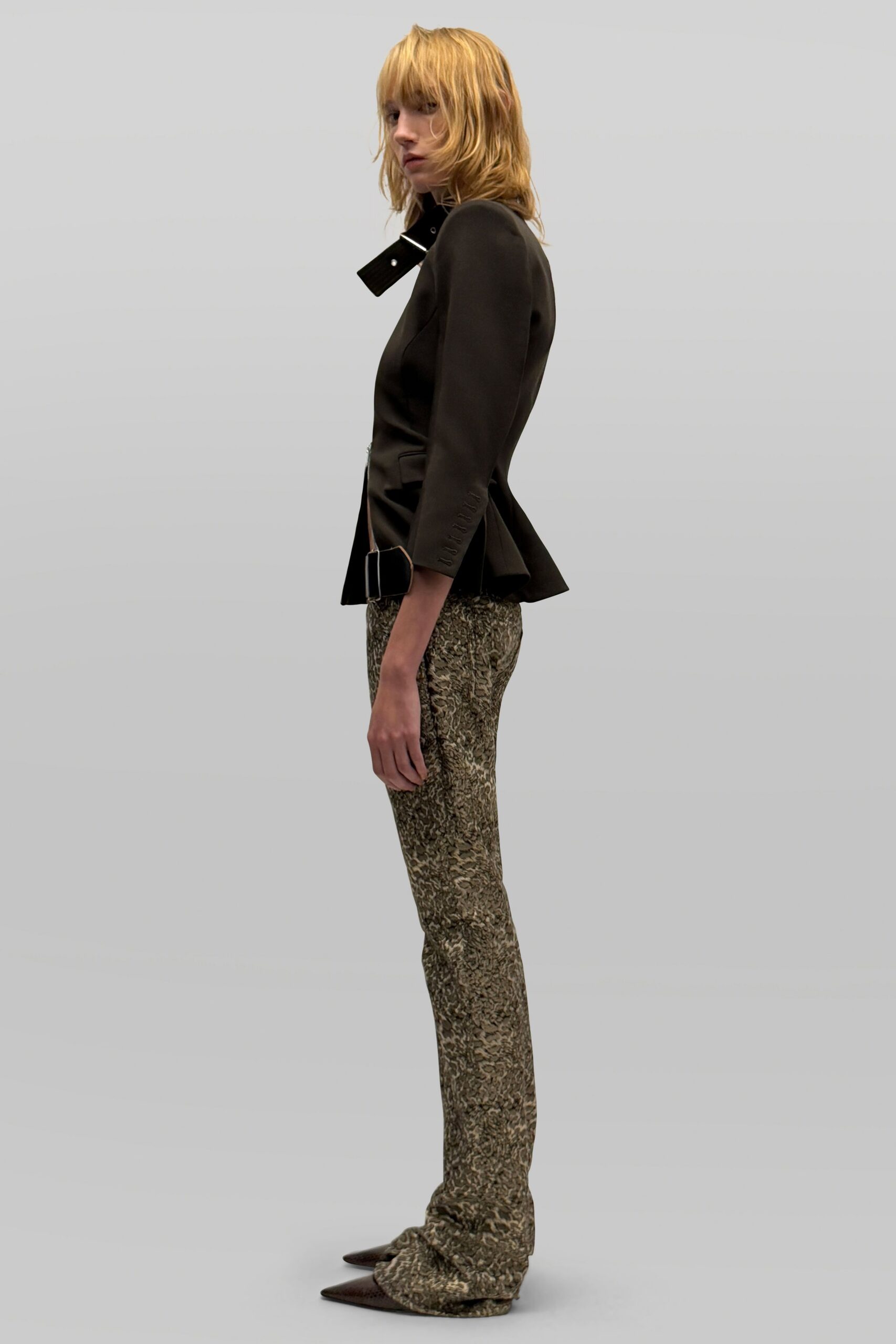 Black tailored jacket with a high tab collar and oversized silver buckles. Slim mottled jacquard trousers, pooling slightly over the foot. Dark pointed toes peeking from the hem. For crossing a hotel lobby before anyone has changed out of dinner clothes.