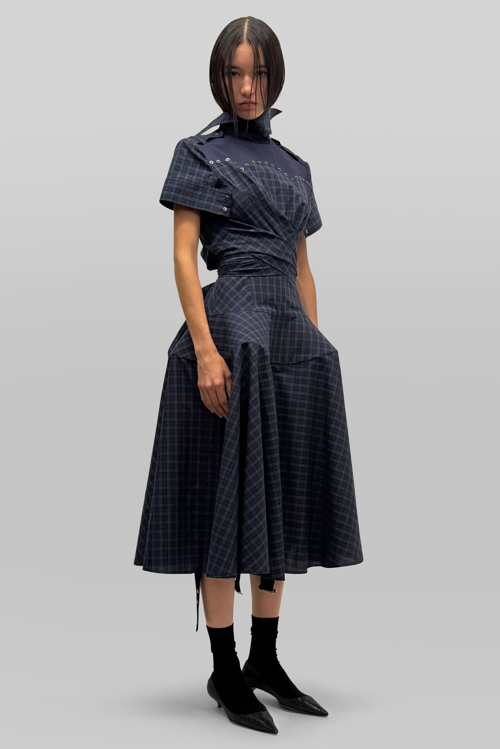 Navy plaid dress with a wrapped bodice, eyelet-trimmed yoke and popped funnel collar. Black ankle socks. Black sock pumps with a reptile-textured pointed cap. Waiting under the station clock after missing the earlier train.