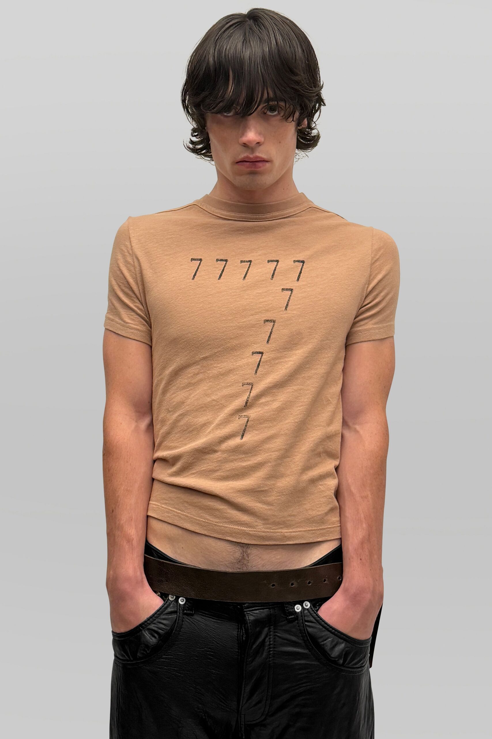Tan fitted jersey tee, black sevens stamped in a descending line. Wide brown leather belt riding low, extra holes marching across. Black leather trousers with inset pocket panels and a wrinkled shine. Waiting at a roadside casino before noon, machines already ringing.