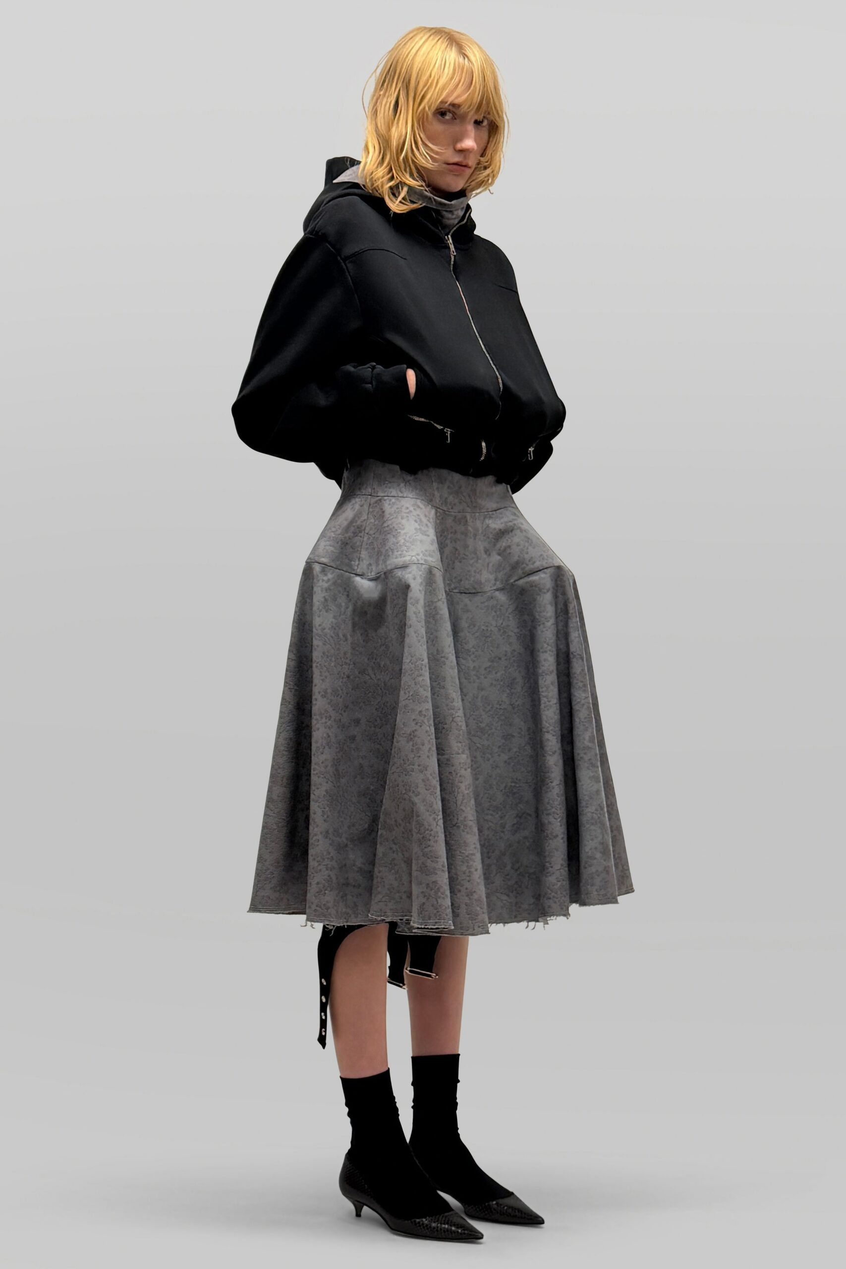 Black cropped hoodie with a silver zip and thick cuffed sleeves. Gray jersey turtleneck peeking close at the throat. Mottled dove-gray skirt cut full, hem left slightly raw. Black sock boots with pointed python-effect toes. Waiting out light rain under a museum overhang after the doors shut.