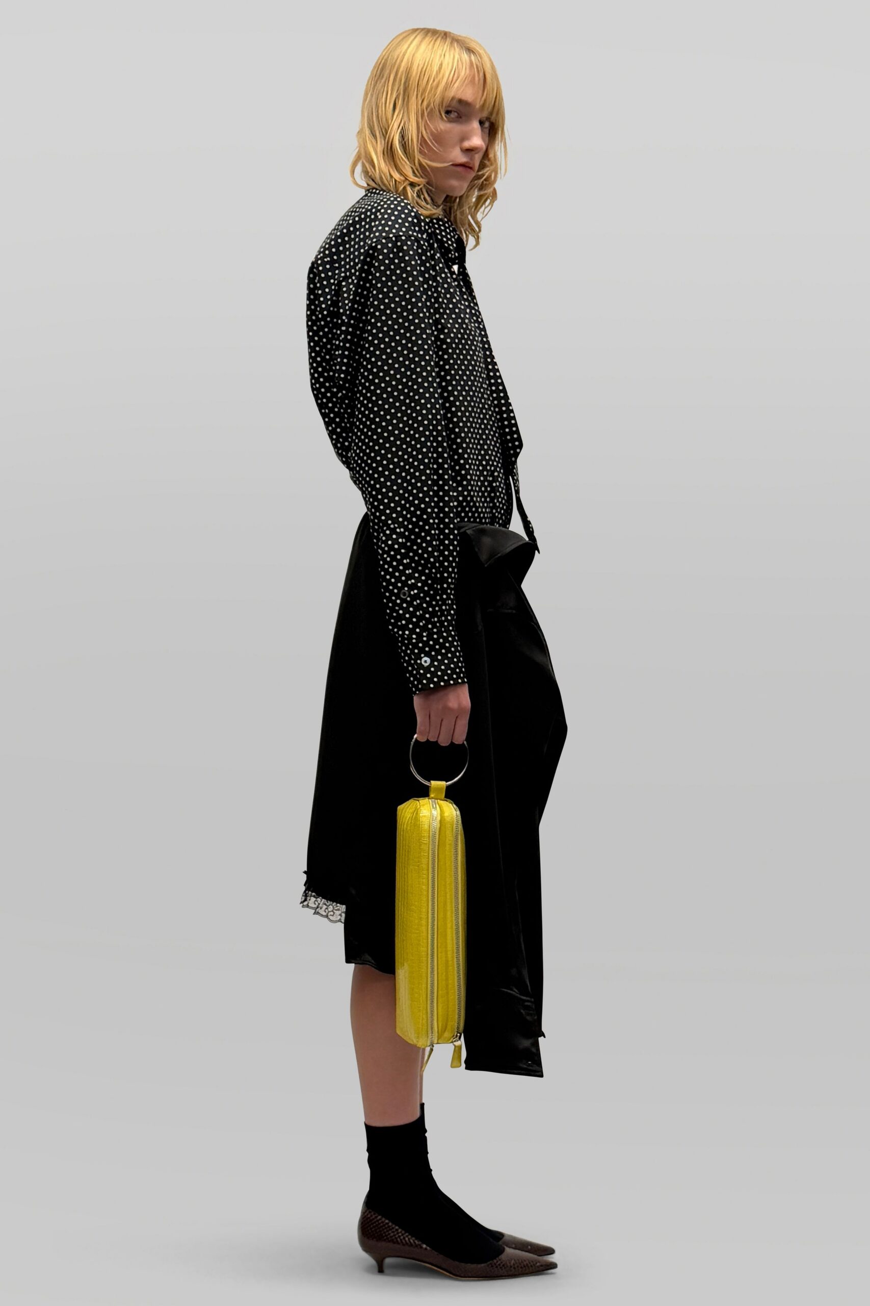 Black blouse scattered with pin-dot white spots, tie hanging loose. Black wrap skirt with a sliver of lace peeking below. Canary case bag with a silver ring handle and long zip. Black ankle socks. Brown mesh kitten heels, sharply pointed. Walking to the station with one lemon tart boxed for later.