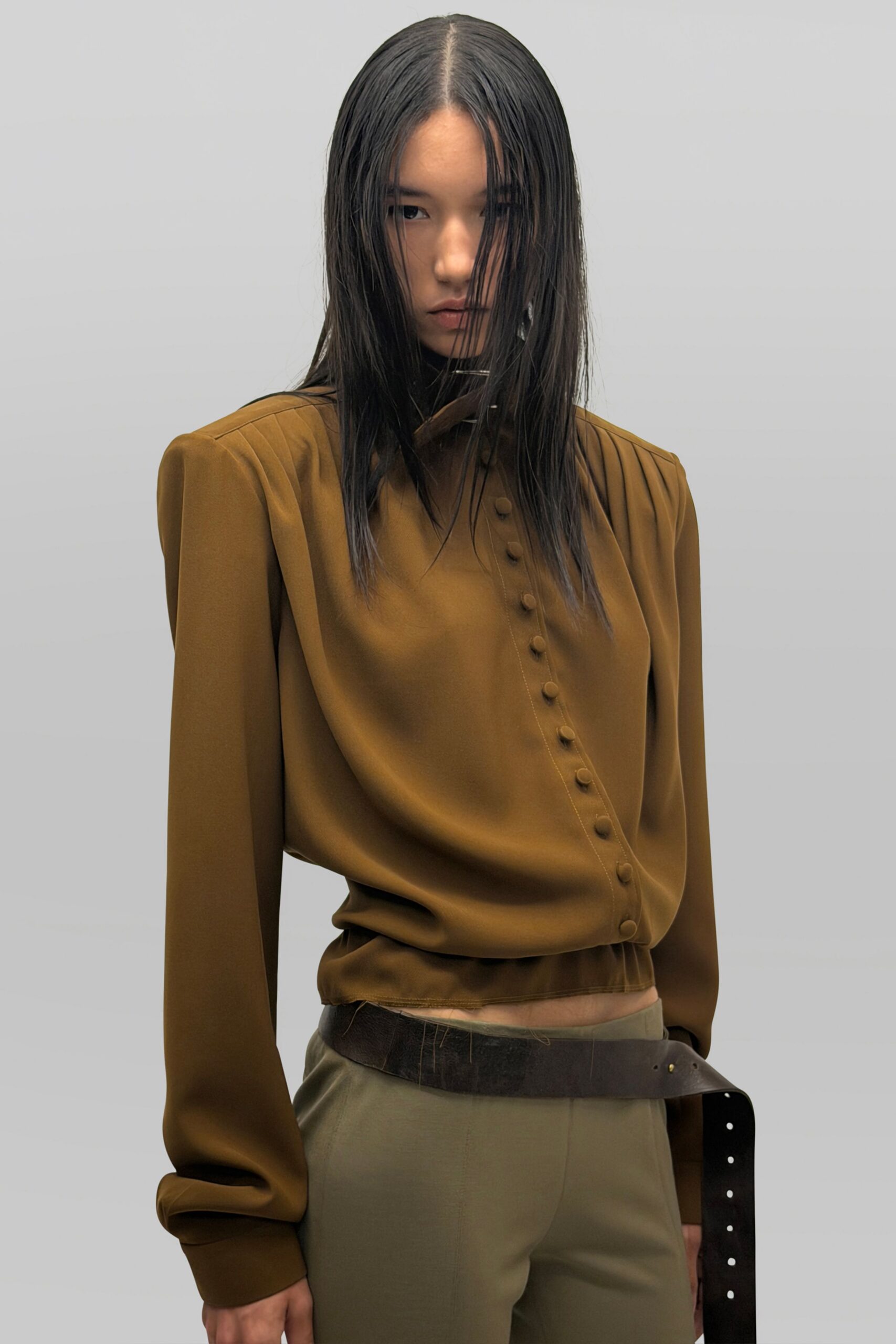 Olive-brown crepe blouse with knife-pleated shoulders and covered buttons set askew. Putty straight trousers with a pressed crease and low slung rise. Wide black leather belt, loose tail punched with bright eyelets. For the last hour in a municipal office after the radiators kicked on.