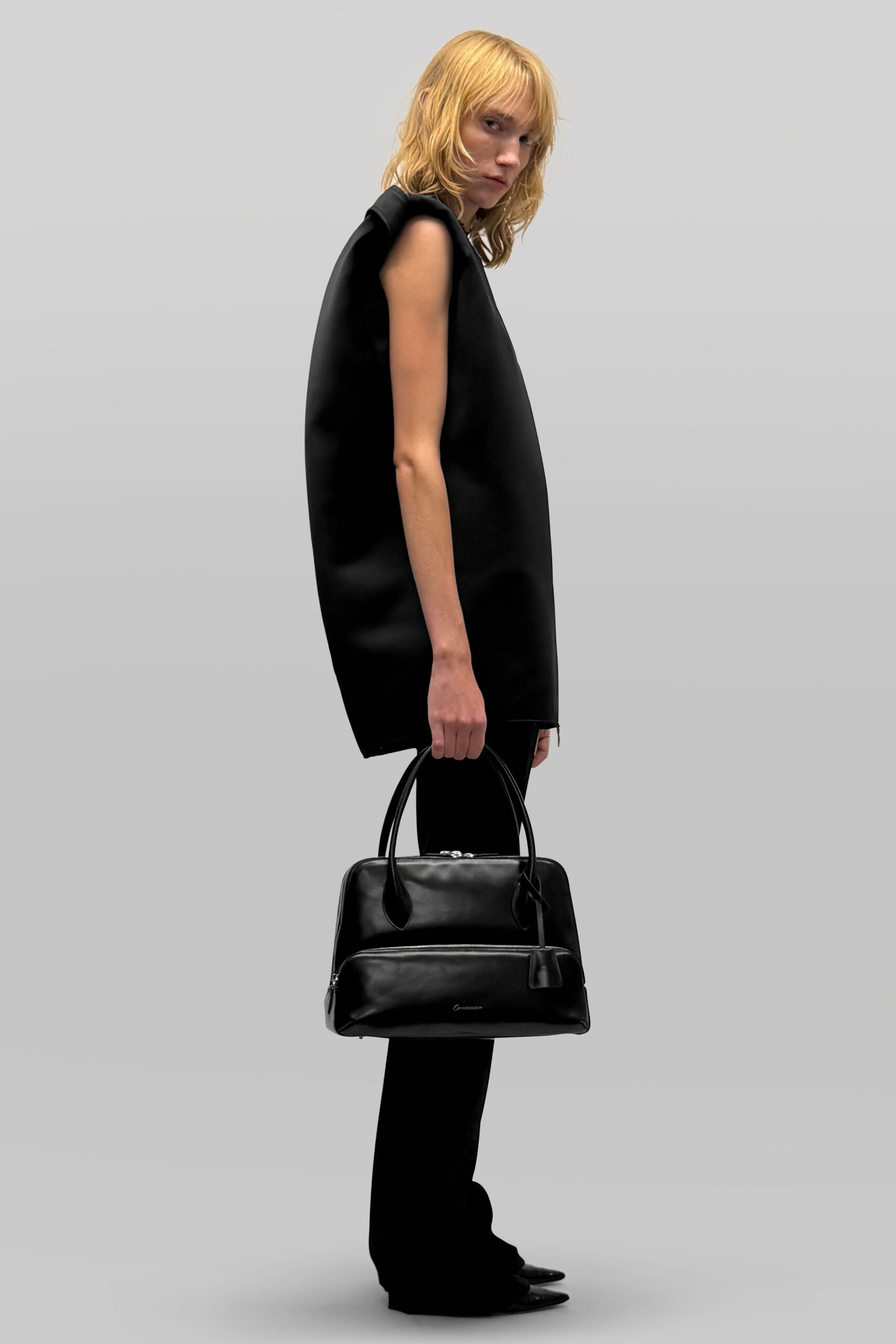 Black structured tunic, sleeveless and cocooned away from the body. Black flared trousers pooling lightly over pointed shoes. Soft black leather holdall with a bulging front zip pocket. For crossing a windswept forecourt with hotel keys and nowhere urgent to be.