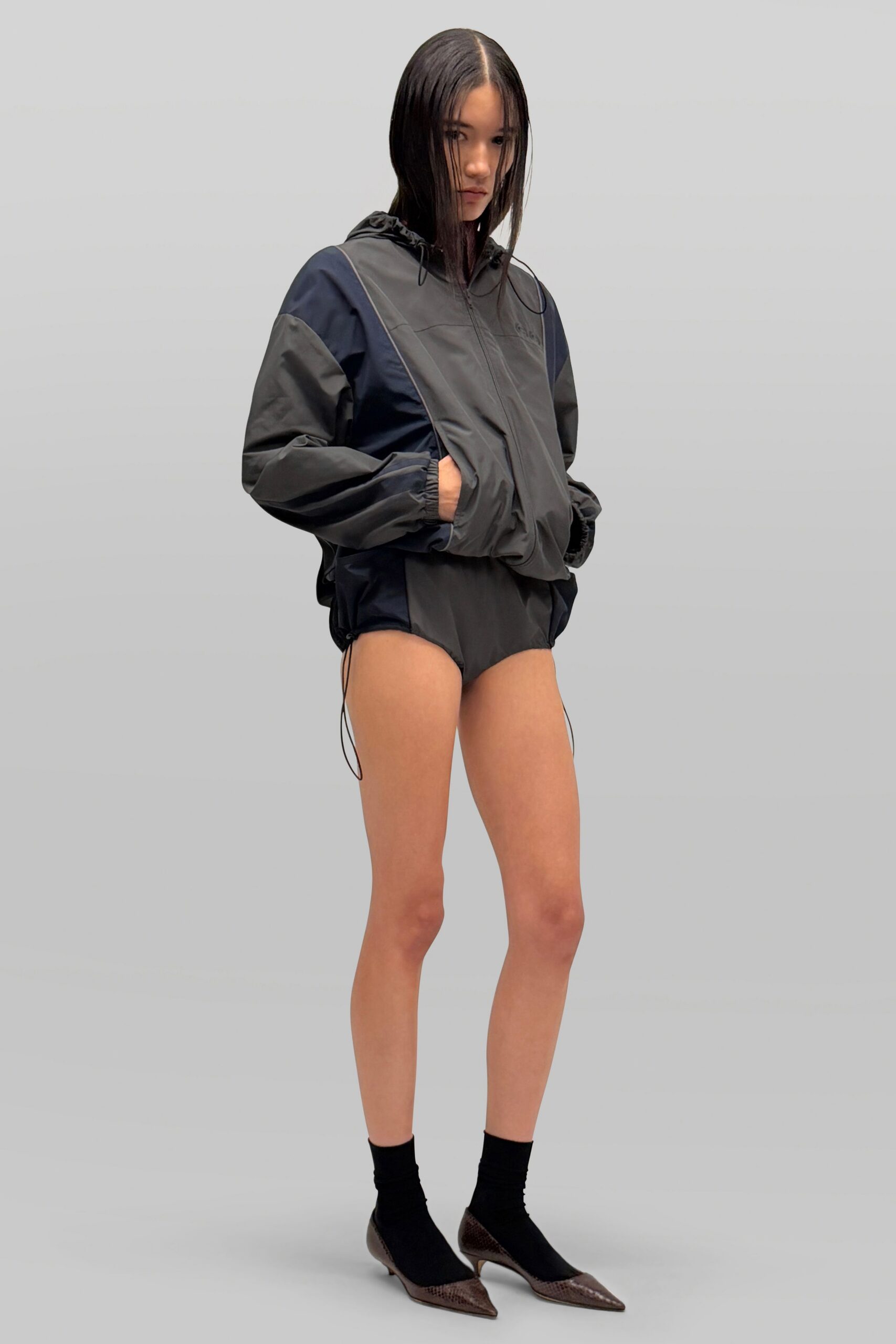 Slate and navy nylon windbreaker, diagonal paneling and bungee cords hanging loose. High-cut black briefs in the same crisp shell. Black ankle socks. Brown python pumps with needle toes and a small metal heel. For buying bottled water and batteries before the storm reaches town.