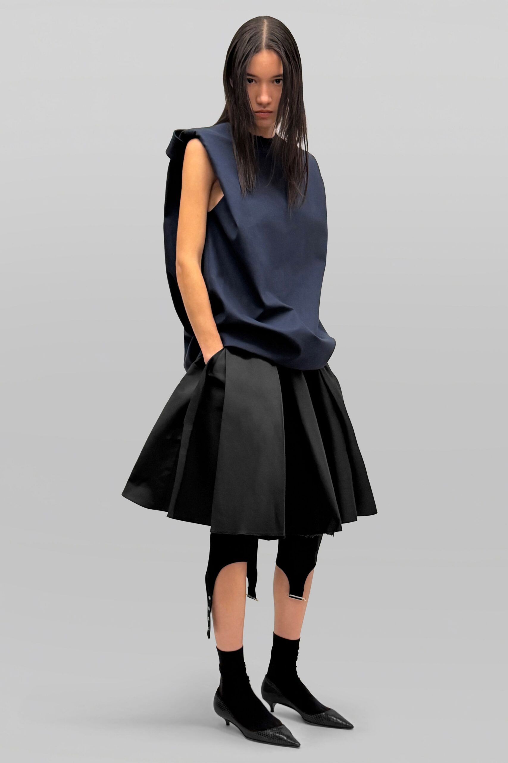 Navy structured shell top, armholes cut deep and standing off the body. Black flared skirt in crisp cloth, one hem corner left fraying. Black stirrup leggings with silver bars at the calf cutouts. Black sock boots with pointed reptile-embossed caps. Crossing an empty museum atrium before the guards unlock the side doors.