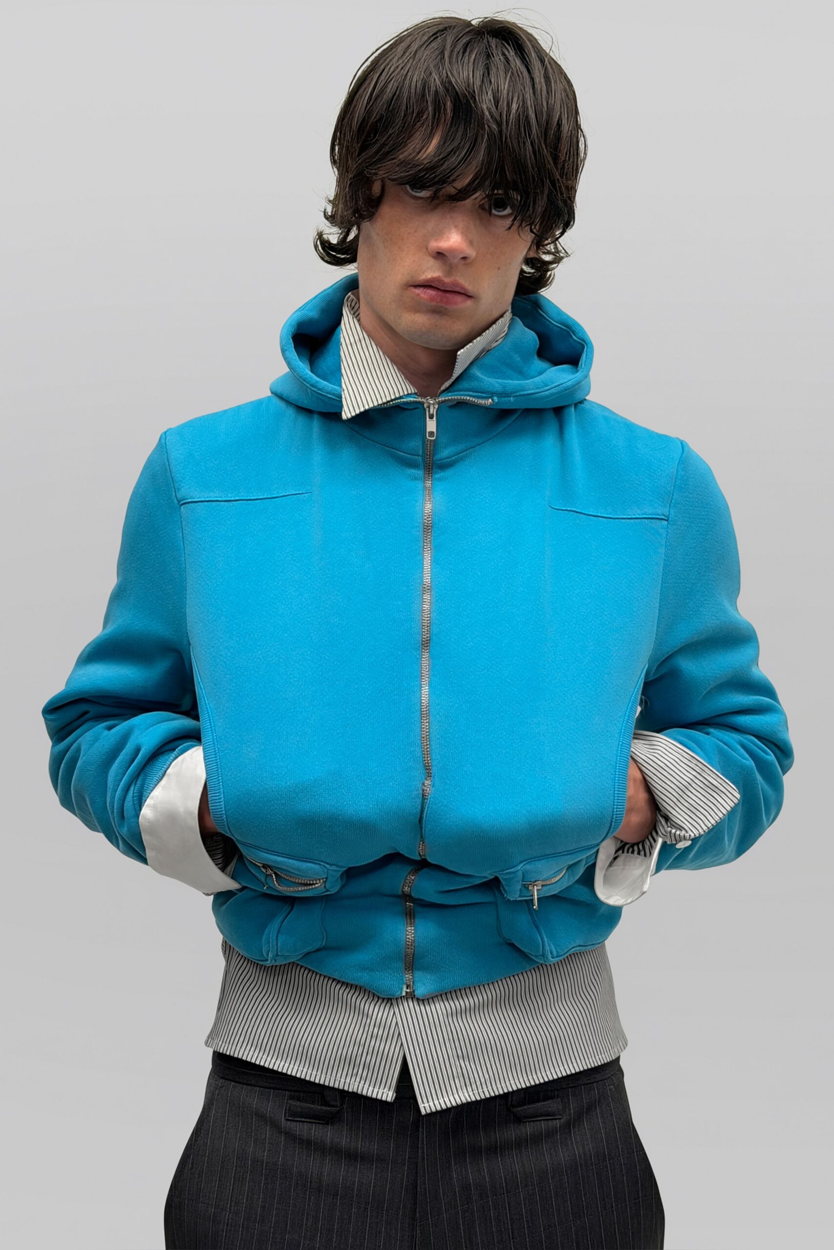 Turquoise bonded jersey hoodie with a second zippered layer bulging at the chest. Grey banker-stripe shirt, stiff collar and long white cuffs jutting out. Charcoal pinstripe trousers, pleated front just visible below the shirttail. Office stairwell smoke break, badge still clipped to your belt.