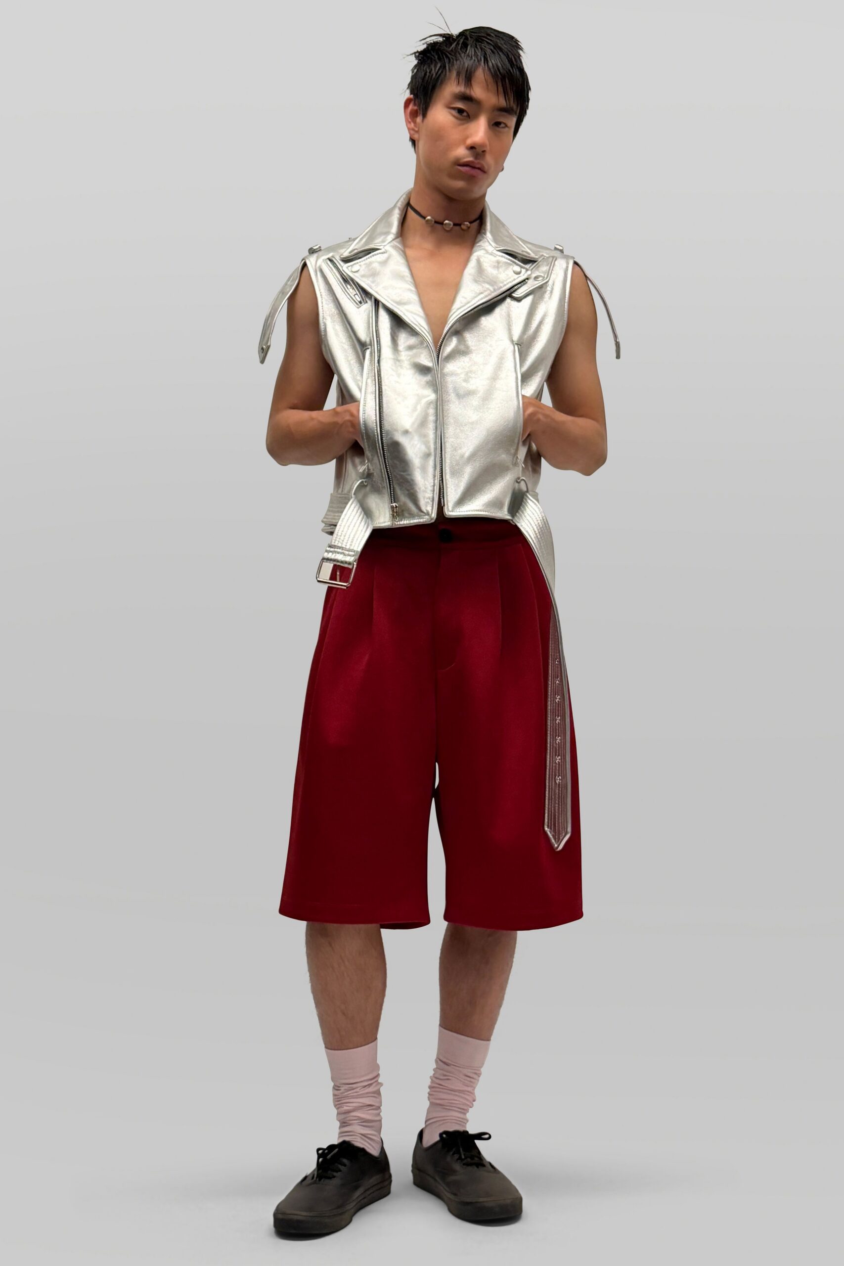 Silver metallic biker vest with dropped armholes and long loose belt straps. Cherry red bermuda shorts cut wide through the leg. Shell bead choker. Pale pink slouched socks. Black lace-up shoes, dull at the toe. For the walk from beach arcade to a bar with sand still on the floor.