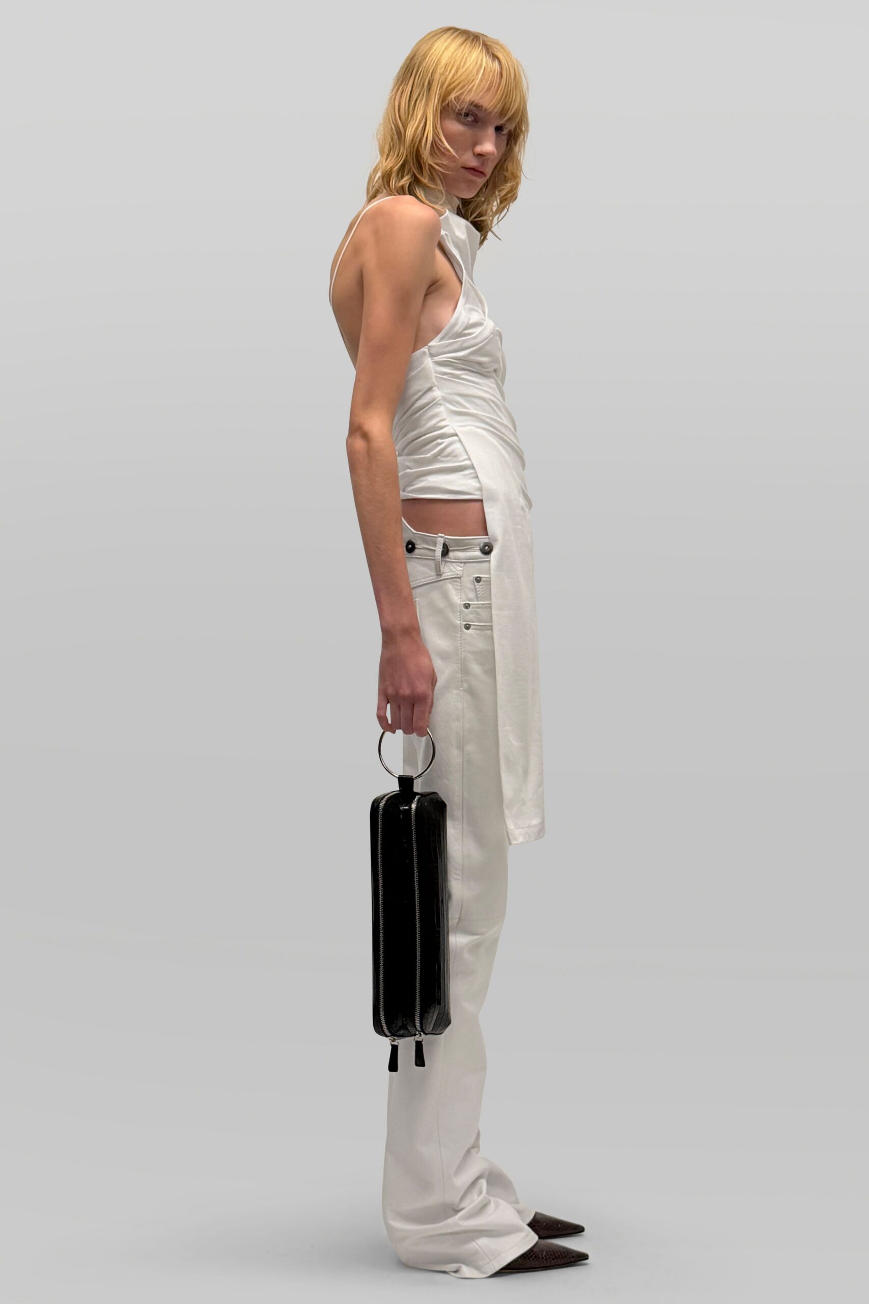 Pearly white satin top, ruched through the waist with a long hanging panel. Ivory low-rise trousers with tabbed waistband and two dark metal studs. Black rectangular case bag on a round silver ring handle. Dark pointed pumps in a fine reptile emboss. For checking into a hotel after midnight with only one hard case.