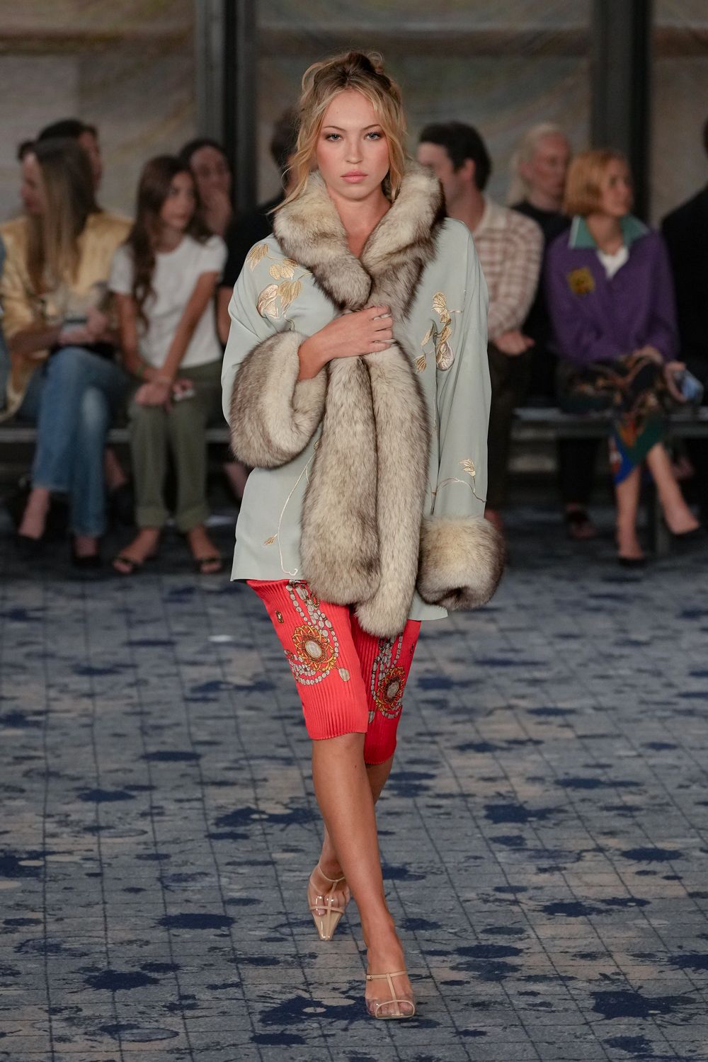 Powder-blue silk coat embroidered with pale gold leaves, huge silvered-fur collar and cuffs. Coral ribbed pencil skirt, jeweled medallion print wrapping one thigh. Nude patent sandals with clear strap across the toes. Hotel corridor at noon, still wrapped in last night's opera coat.