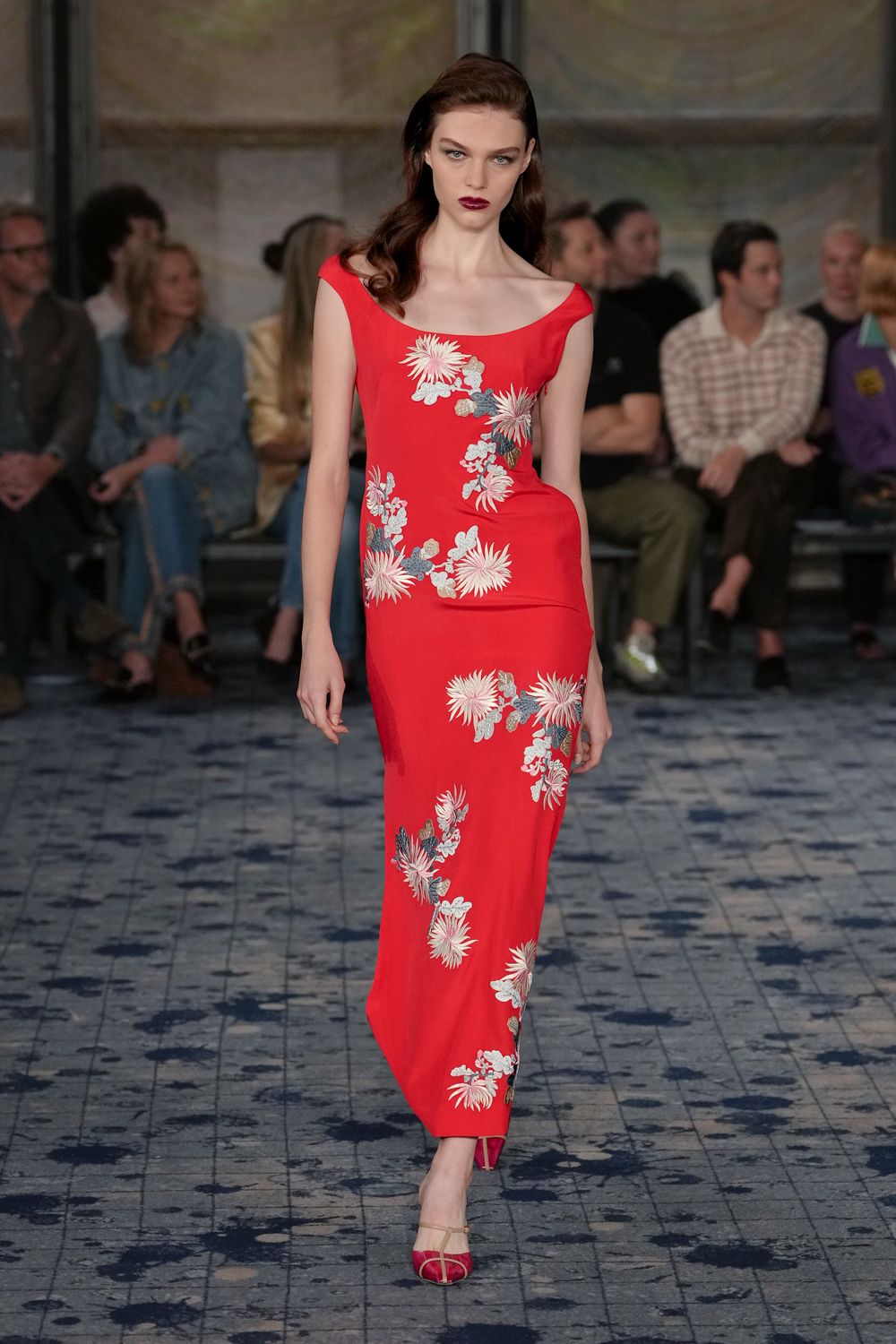 Scarlet column gown with cap sleeves and pale chrysanthemum embroidery climbing diagonally. Red satin peep-toe heels with a clear strap across the instep. Crossing a hotel lobby during wedding season, flowers already dropping.