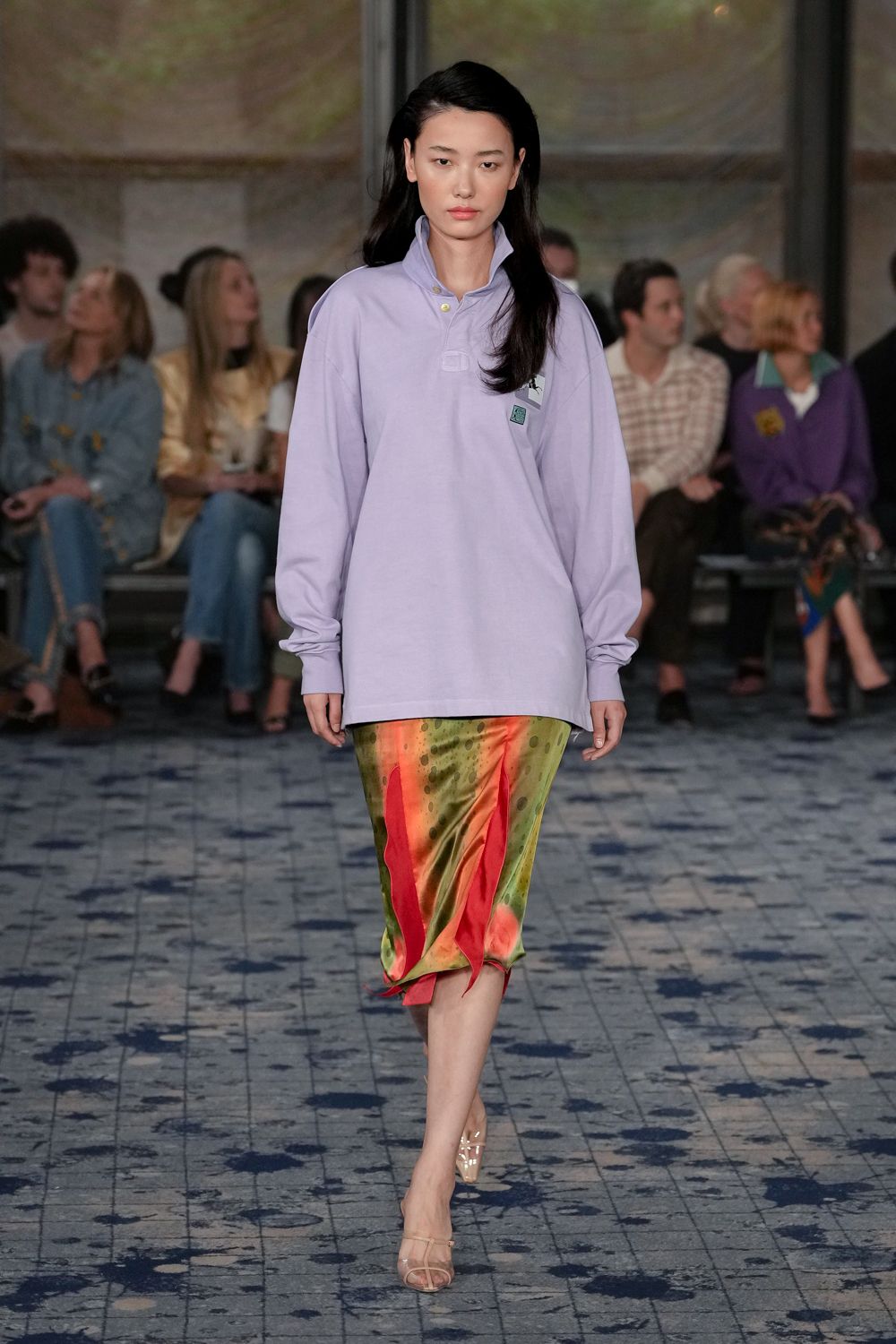 Oversized lilac cotton pique polo, collar popped and cuffs pooling slightly. Bias-cut satin midi printed in olive and coral, pointed godets flicking at the hem. Clear strap heeled sandals with a glossy nude footbed. Walking to a hotel terrace lunch while the fountain spray keeps catching your calves.