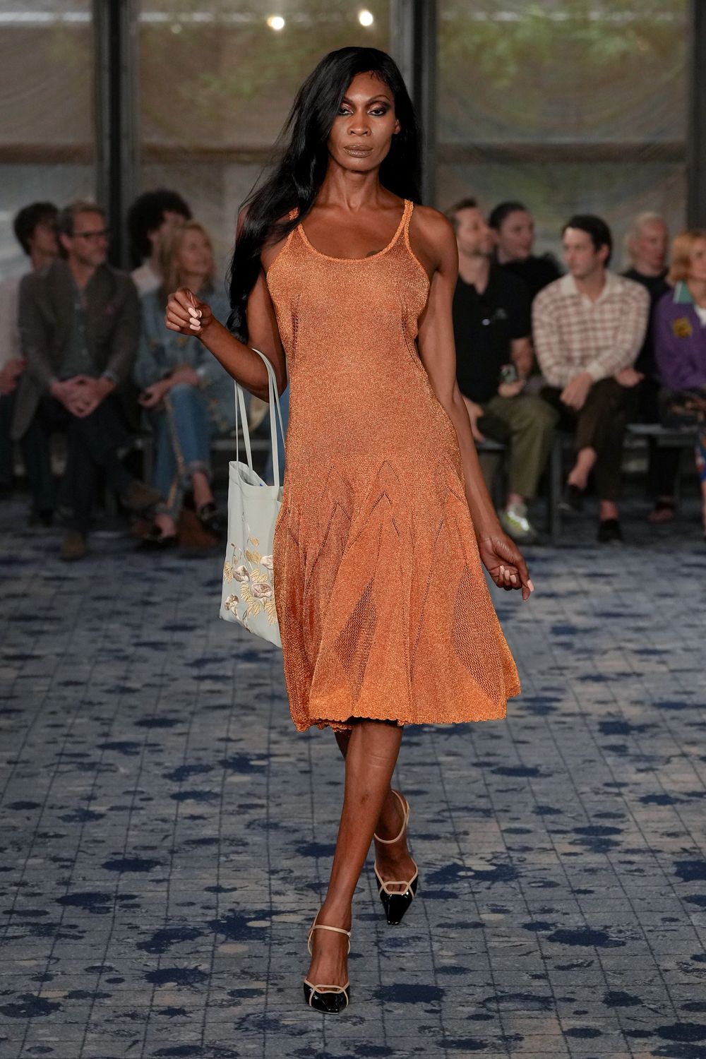 Copper open-knit slip dress, dropped panels flaring from zigzag seams. Pale blue tote with a faded floral print near the base. Cream slingbacks with black patent cap toes and tiny buckle straps. Hotel courtyard after rain, carrying back cut flowers and somebody else's book.