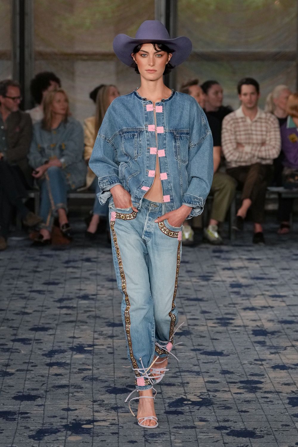 Periwinkle felt cowboy hat with an exaggerated rolled brim. Washed blue denim jacket, raw neckline and pink frog closures. Pale jeans trimmed with geometric braid down the outseam and pockets. White lace-up sandals, cords snaking messily around the ankles. County fair heat, somewhere between the livestock pens and the beer tent.