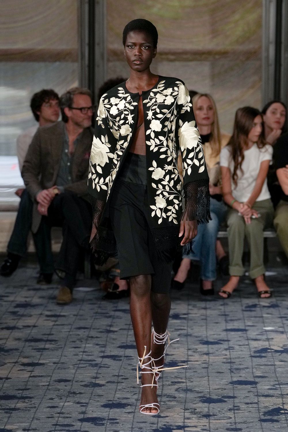 Black coat covered in ivory floral embroidery, mesh fringe at cuffs and hem. Sheer brown mesh panel meeting at a narrow front clasp. Black wrap skirt with a slanted overlap cutting across the thigh. White lace-up sandals, ties jutting loose at the ankles. Crossing a tiled courtyard just after the wedding meal has thinned out.