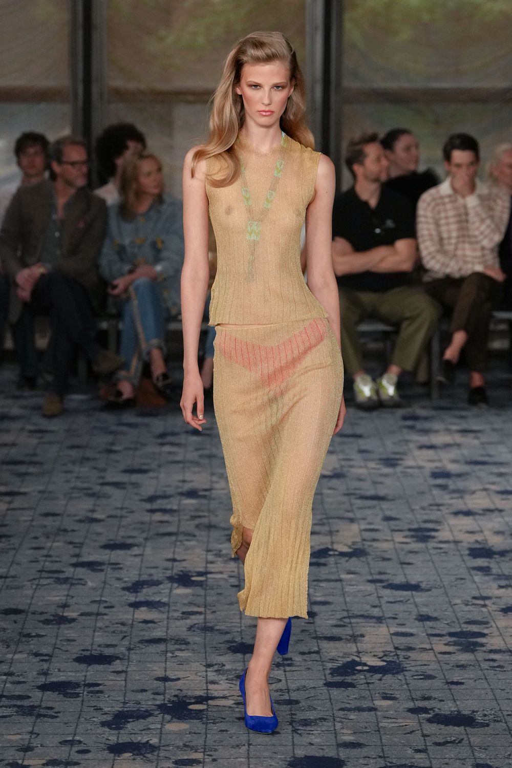 Fine gold ribbed knit, sheer enough to show the chest beneath. Matching ribbed skirt with a coral chevron panel slung across the hips. Electric blue suede pumps. Walking into a hotel atrium while the fountain is still being switched on.