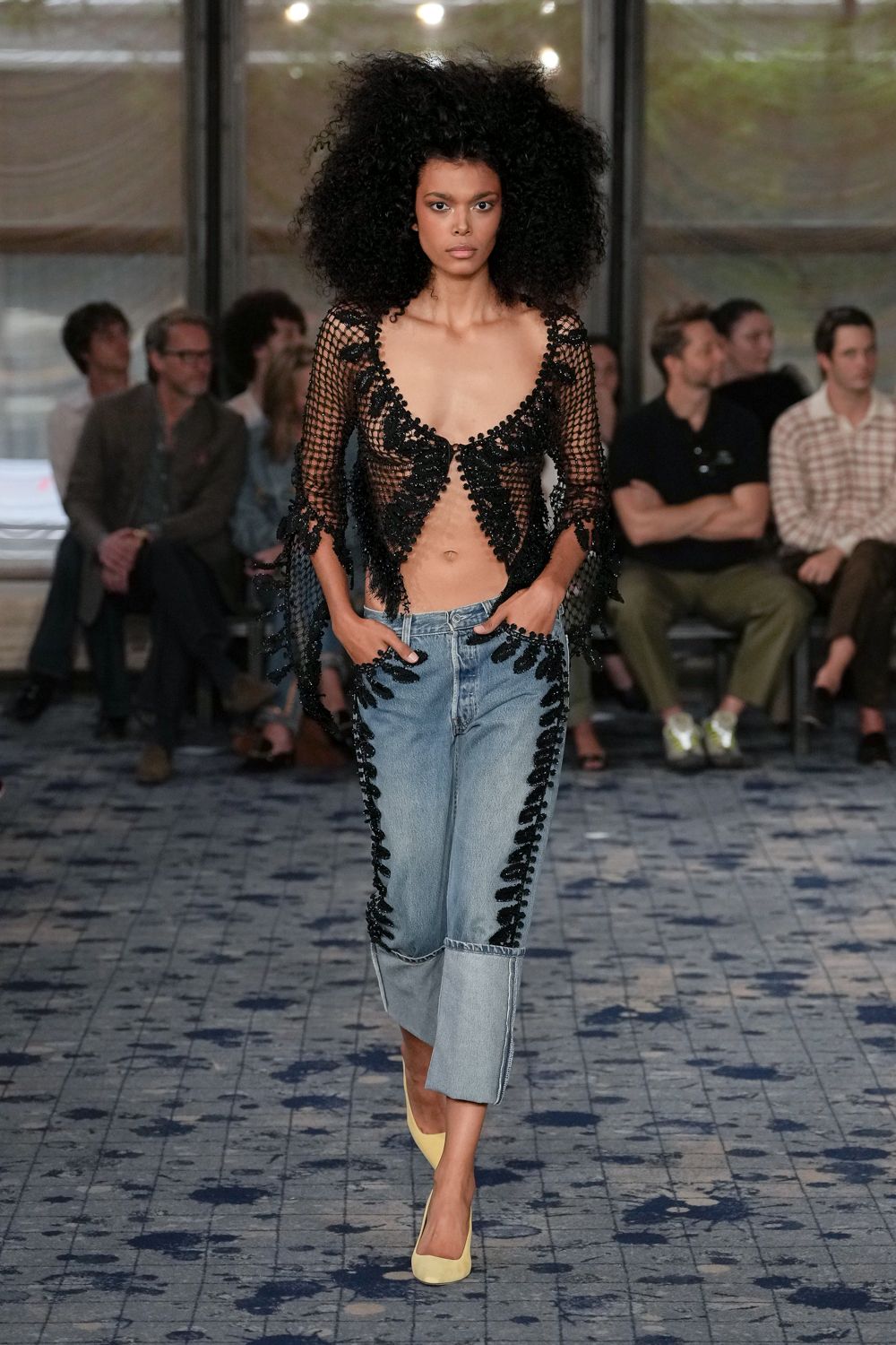 Black openwork mesh top edged with dense beaded appliqué, split at the waist. Washed blue cropped jeans traced with black beaded scrolls, deep turned-back cuffs. Butter-yellow pumps, plain and closed-toe. Crossing a hotel atrium after rain, still half dressed for somewhere warmer.
