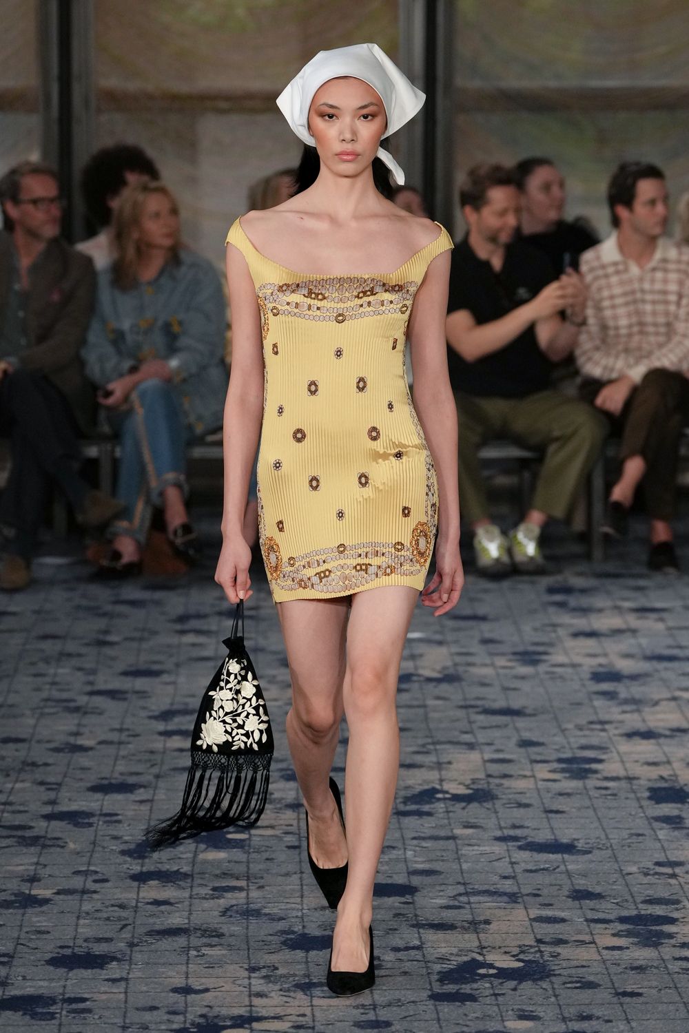White folded headscarf tied low behind one ear. Butter yellow ribbed knit mini, chain-and-medallion print skimming the neckline. Black floral tapestry pouch with long swaying fringe. Black suede pumps, sharply cut at the vamp. Crossing a hotel courtyard after breakfast, room key still in hand.