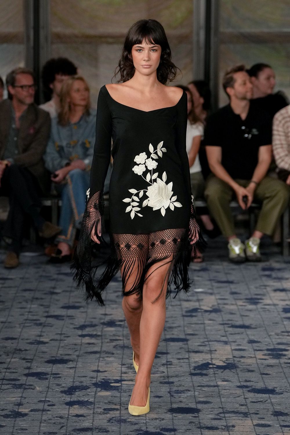 Black boat-neck mini dress with ivory floral applique climbing the hip. Fishnet sleeve ends spilling into long black fringe. Open net hem dotted with small rosettes above swinging strands. Buttercream pumps with a rounded topline. Hotel terrace wedding, wind catching the fringe before the speeches start.