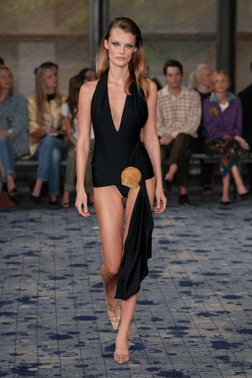 Black jersey halter dress, plunging to the navel with a shell clasp at the hip. High-cut black briefs exposed beneath the wrapped drape. Nude ankle-strap sandals with clear pointed shields over the toes. Pool terrace after sunset, skin still salted and the tiles cooling off.