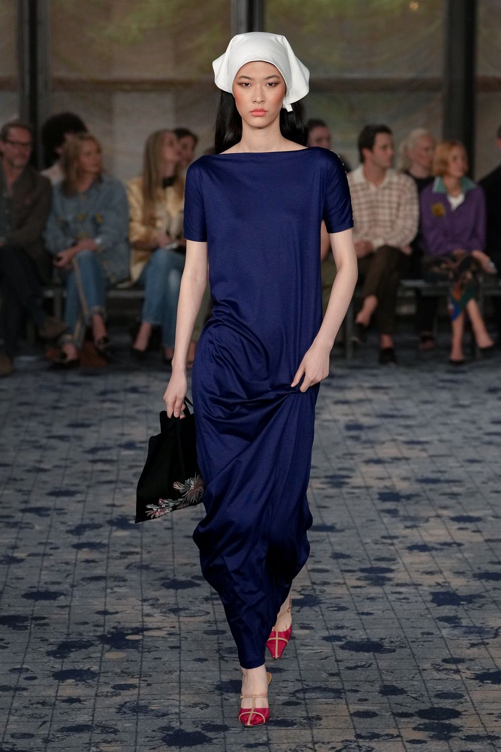 White cotton headscarf folded into a soft squared cap. Ink-blue jersey column dress, bateau neck and ruching caught at one hip. Black fabric tote with a pale feathered spray at the corner. Raspberry strappy sandals with a narrow beige T-bar. Crossing a tiled courtyard just after the fountain has been turned off.