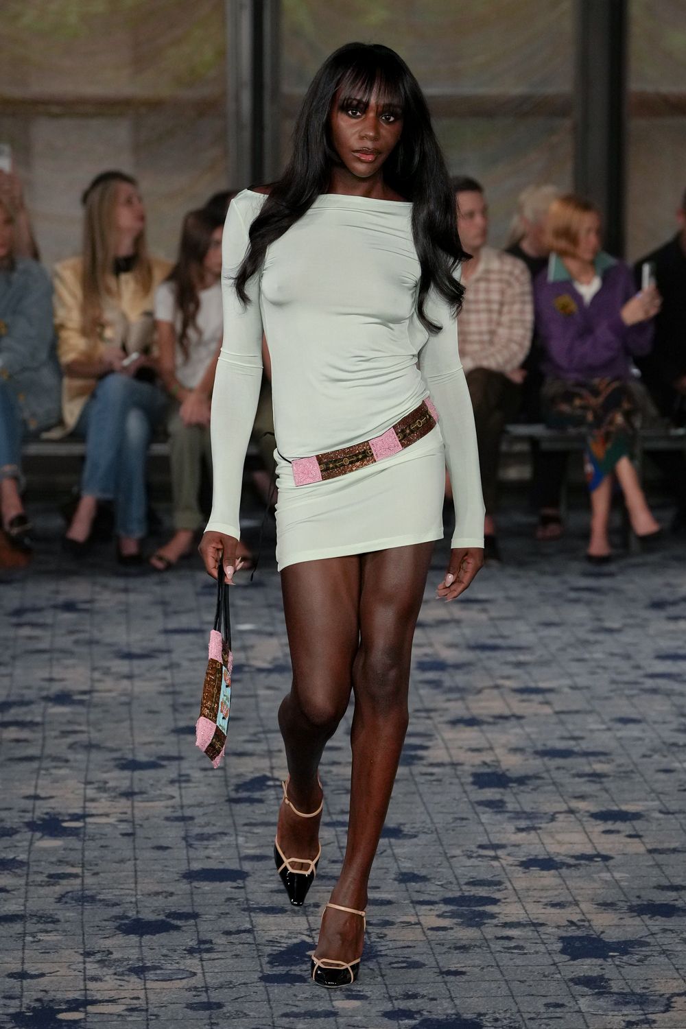 Pale mint stretch mini dress, bateau neckline and one sliced shoulder seam. Low-slung patchwork belt with pink boucle tabs and tiny ring hardware. Slim matching pouch, black cord handles swinging from one hand. Cream slingbacks with glossy black cap toes. Walking back from a motel ice machine in the first light.