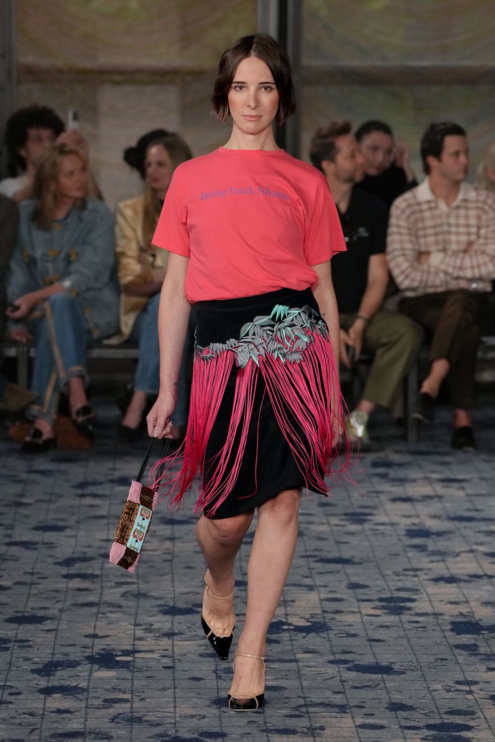 Hot-pink cotton jersey tee with blue slogan text across the chest. Black skirt draped with grey leaf appliques and long pink fringe. Beaded box bag with glittery pink ends and a black strap. Black patent cap toes with nude mesh and slim ankle straps. Walking into a charity lunch where the raffle prizes are already gone.