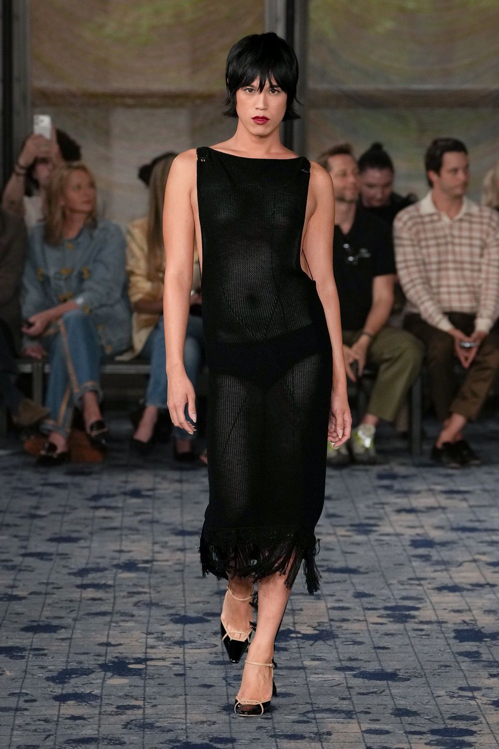 Black open-knit column dress, bateau neck and shredded fringe at the hem. High-cut black briefs visible beneath the loose mesh. Cream slingback heels with black patent cap toes and ankle ties. Walking back from the hotel pool after dark, still half dressed.