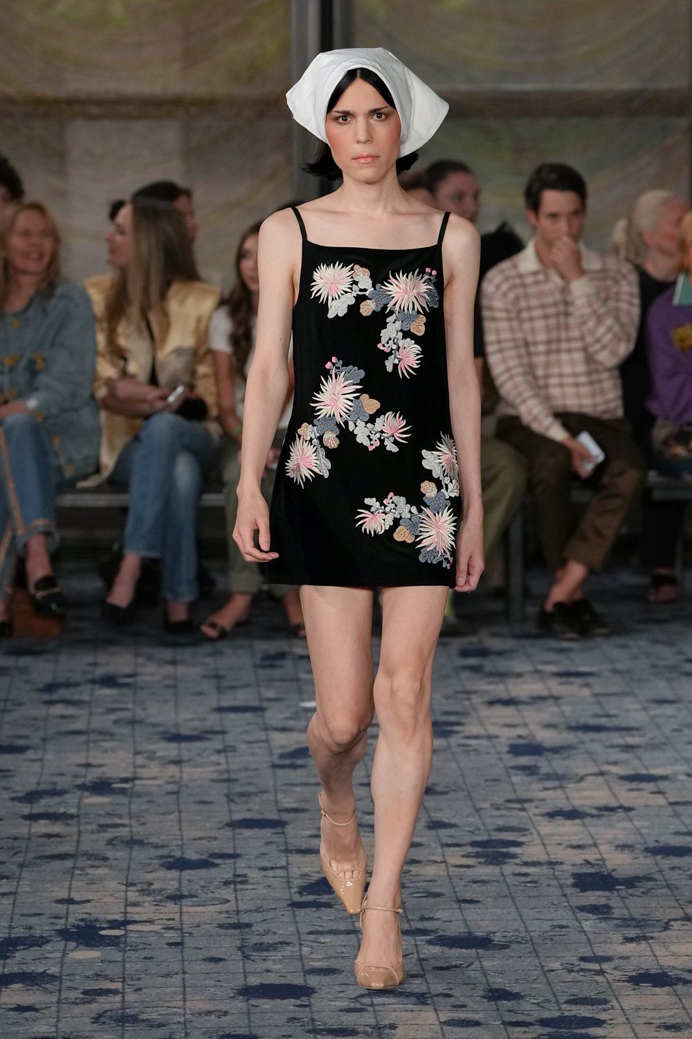 Crisp white headscarf folded into a bonnety wrap around the crown. Black mini slip dress scattered with pale pink chrysanthemum embroidery. Nude patent Mary Jane pumps with a fine instep strap. Church basement wedding, cake already cut and the folding chairs scraped back.