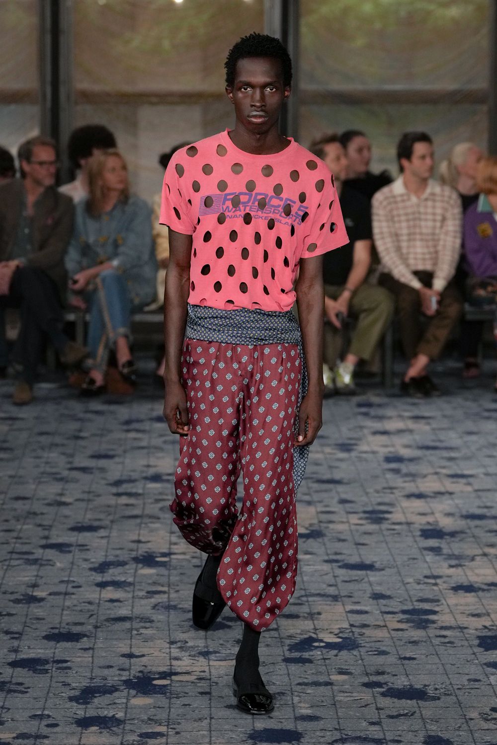 Neon pink tee punched with oval cutouts over black mesh. Blue foulard sash wrapped low, one tail hanging at the hip. Burgundy pajama-cut trousers scattered with tiny medallion motifs. Black sock boots with square patent toes. Walking back from the pool bar before the sunburn starts to sting.