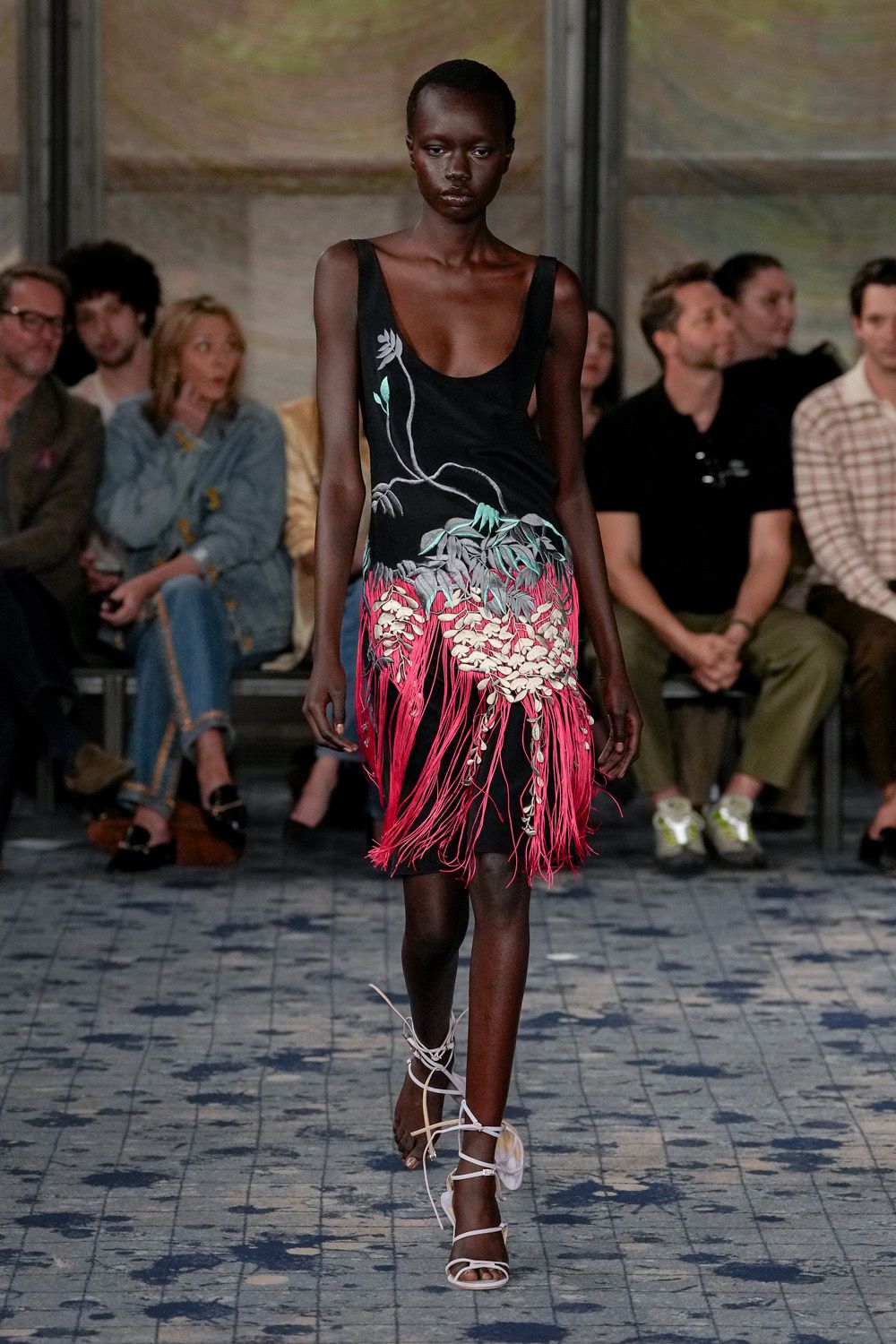 Black slip dress with painted vine blossoms and hot-pink fringe bursting below the hips. White lace-up sandals, a shell-like petal cupping each ankle. Walking the tiled promenade after rain, hotel garden lights coming on.