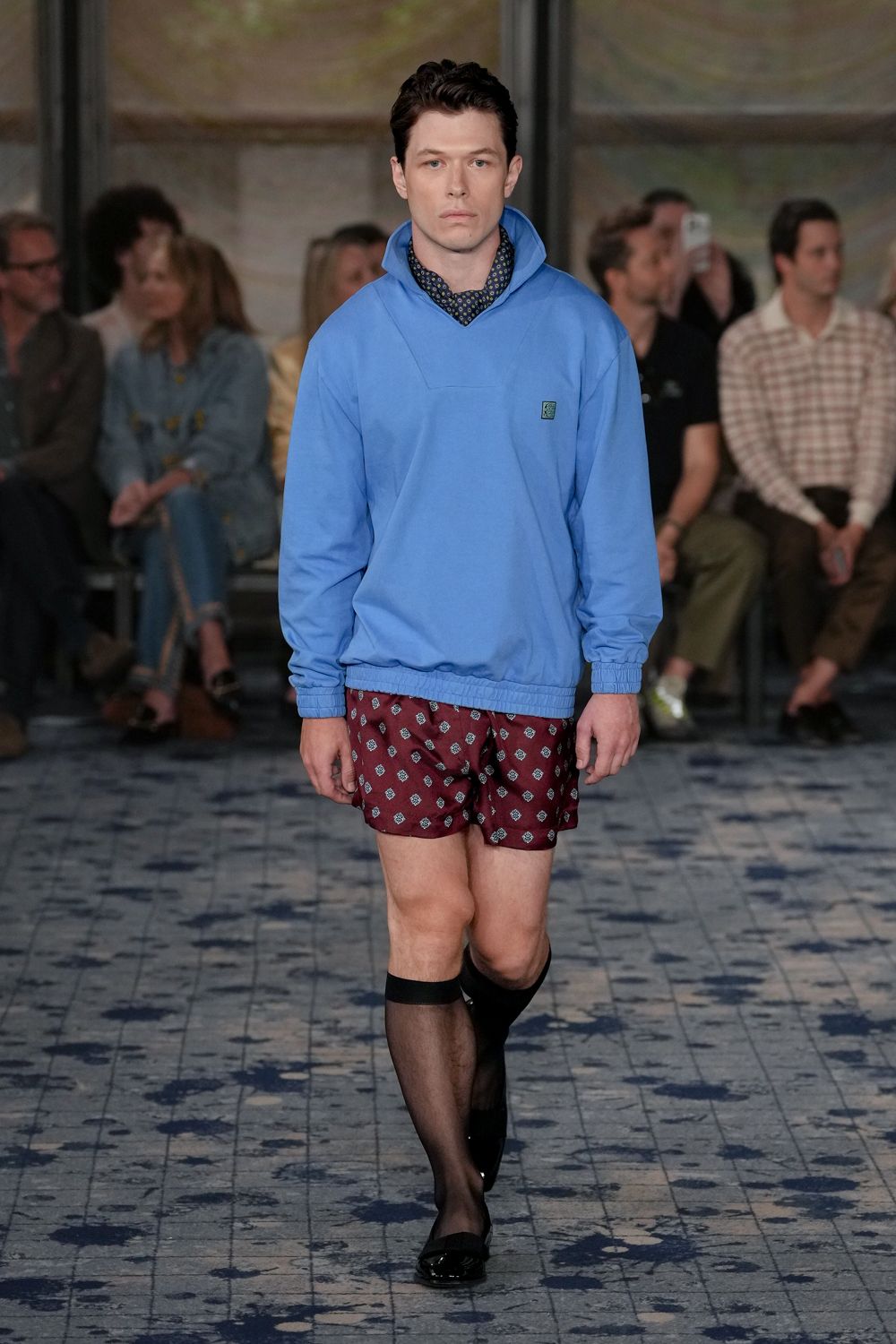 Cornflower blue sweatshirt with a high funnel neck and shirred hem. Navy neckerchief dotted with tiny ochre ring motifs. Burgundy satin shorts scattered with neat foulard diamonds. Sheer black knee-high stockings with opaque bands. Black patent loafers. Hotel terrace at noon, one drink in and nowhere urgent next.