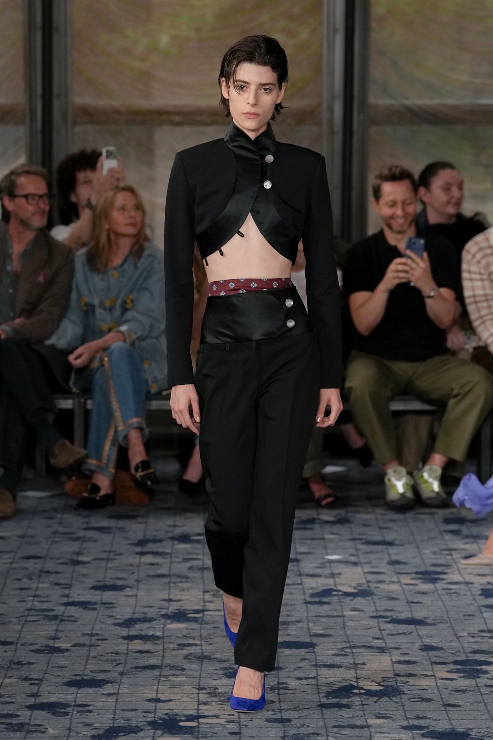 Black satin-faced cropped jacket, curving open over the ribs. Wine jacquard waistband peeking above the cutaway hip. Black trousers with a glossy wrap panel and two silver buttons. Cobalt suede pumps. Whatever you wear to sign papers, then leave by the side entrance.