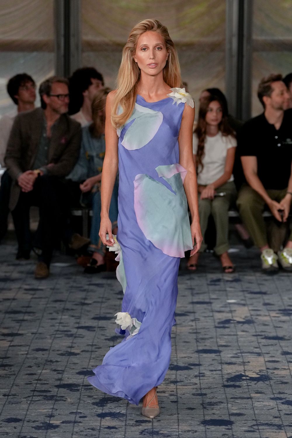 Periwinkle chiffon column gown with translucent petal appliques drifting across it. White organza blossoms pinned at the shoulder, hip, and lower hem. Nude pointed pumps with clear slingback straps. Garden wedding reception after the heat finally breaks.