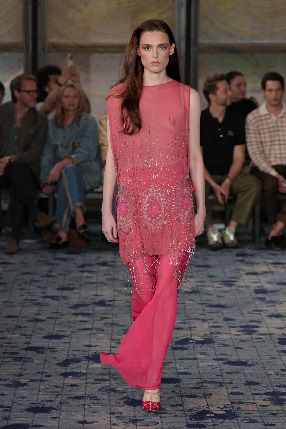 Sheer watermelon tunic, silver bugle-bead fringe and sunburst panels swinging low. Matching chiffon flares, one hem drifting longer across the step. Pink satin sandals with slim metallic ankle straps. Crossing a hotel terrace just before the wedding band starts.