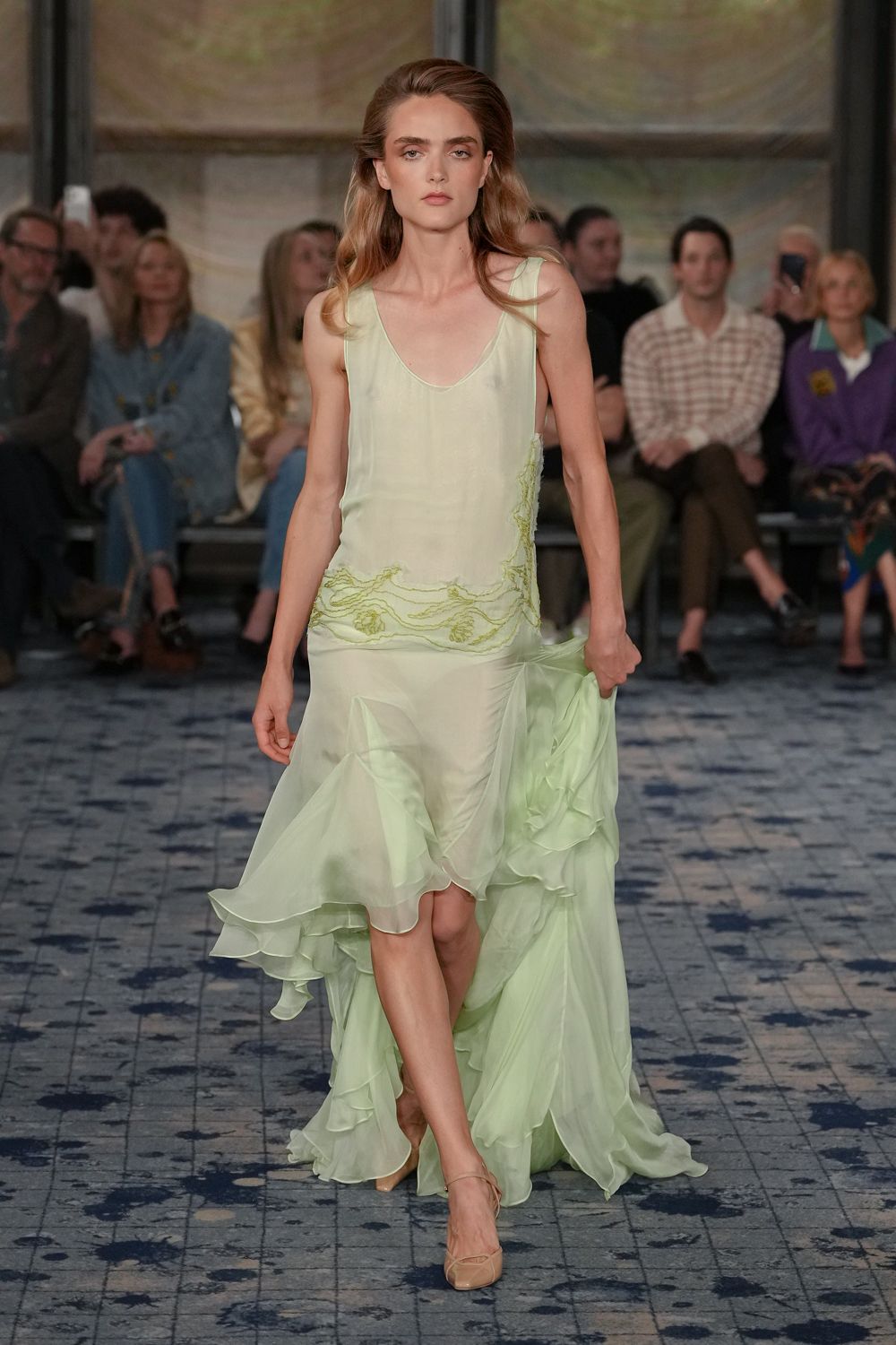 Pale celadon silk slip dress with seaweed-like beading skimming one hip. Sheer chiffon train gathered in the hand, hem breaking into soft ruffles. Nude ankle-strap heels with a rounded, almost shell-like toe. Crossing a hotel terrace just before the garden wedding starts.