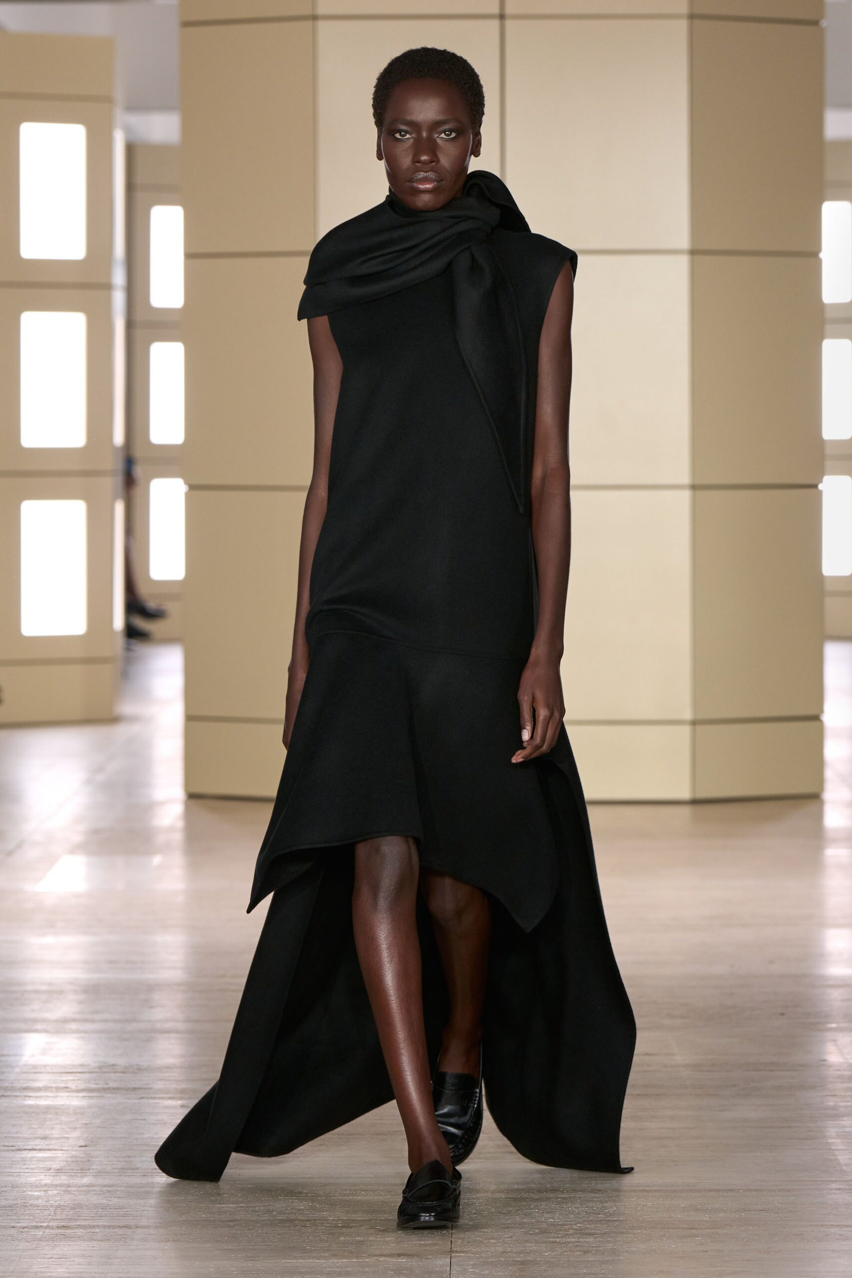 Black sleeveless dress with a wrapped scarf collar and dropped flounce hem. Black leather loafers with a softly squared apron toe. Crossing a silent hotel atrium before anyone else is awake.