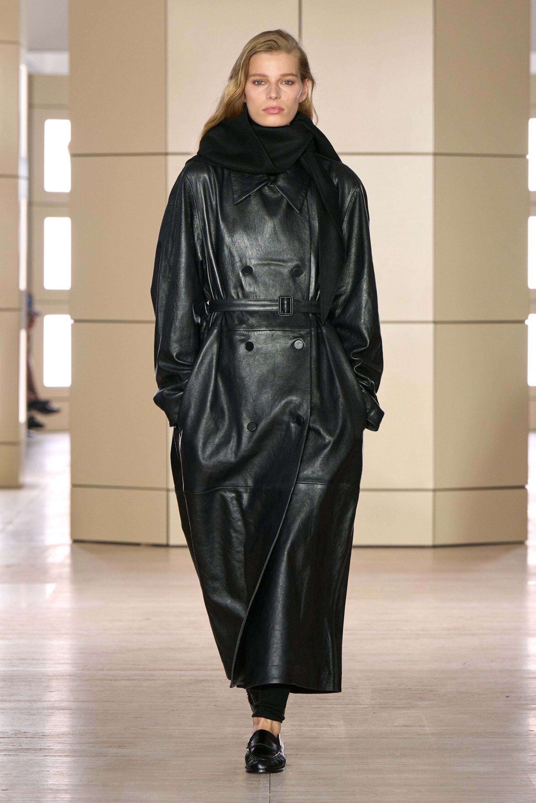 Heavy black wool scarf looped high around the jaw. Glossy black leather trench, double-breasted with a narrow buckled belt. Black leggings disappearing under the long hem. Black penny loafers with a softly squared apron seam. For crossing a silent hotel lobby before anyone else is awake.