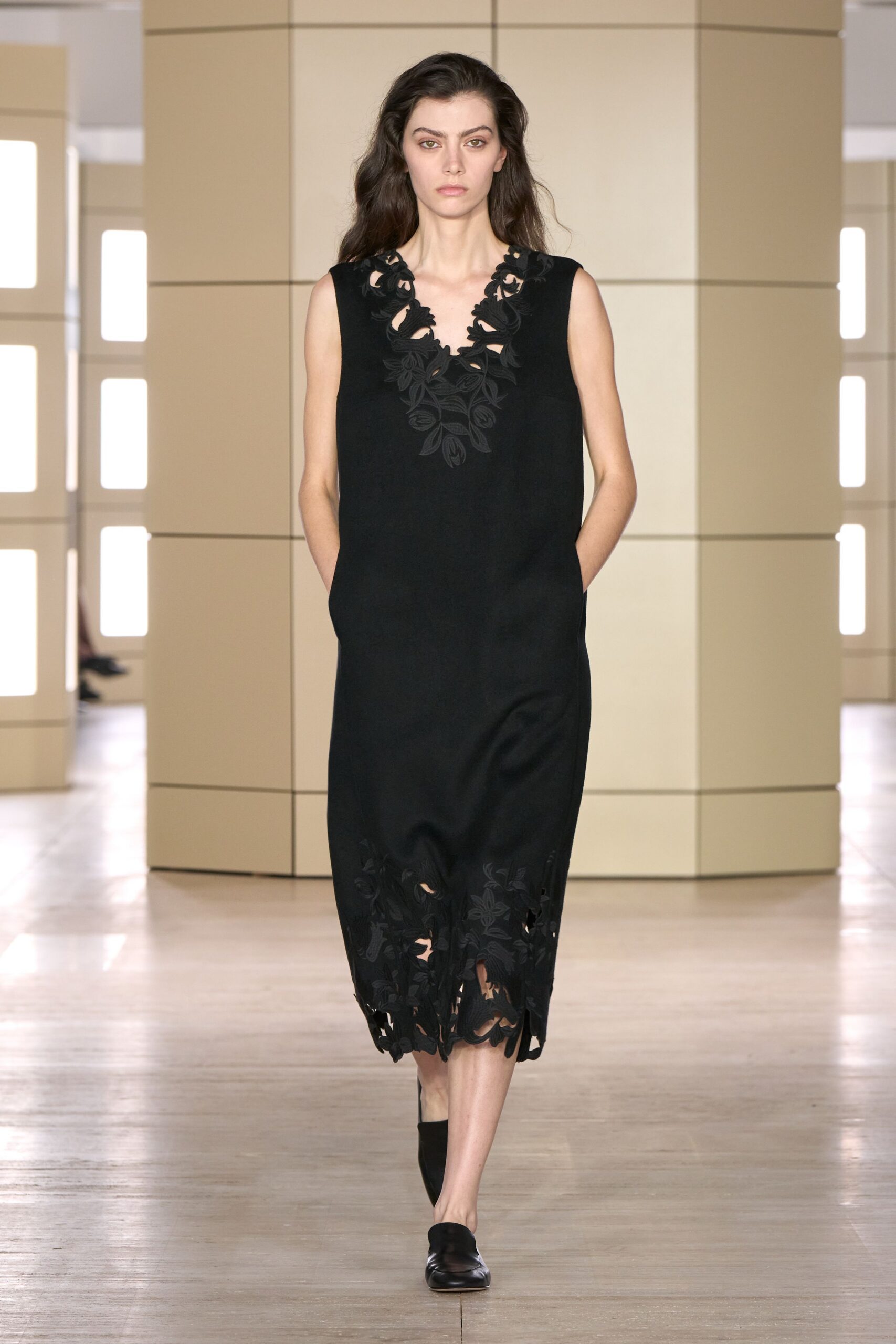 Sleeveless black shift dress with floral cutwork opening the deep V neck and hem. Black leather mules with a squared vamp. Museum atrium at noon, footsteps echoing off pale stone.