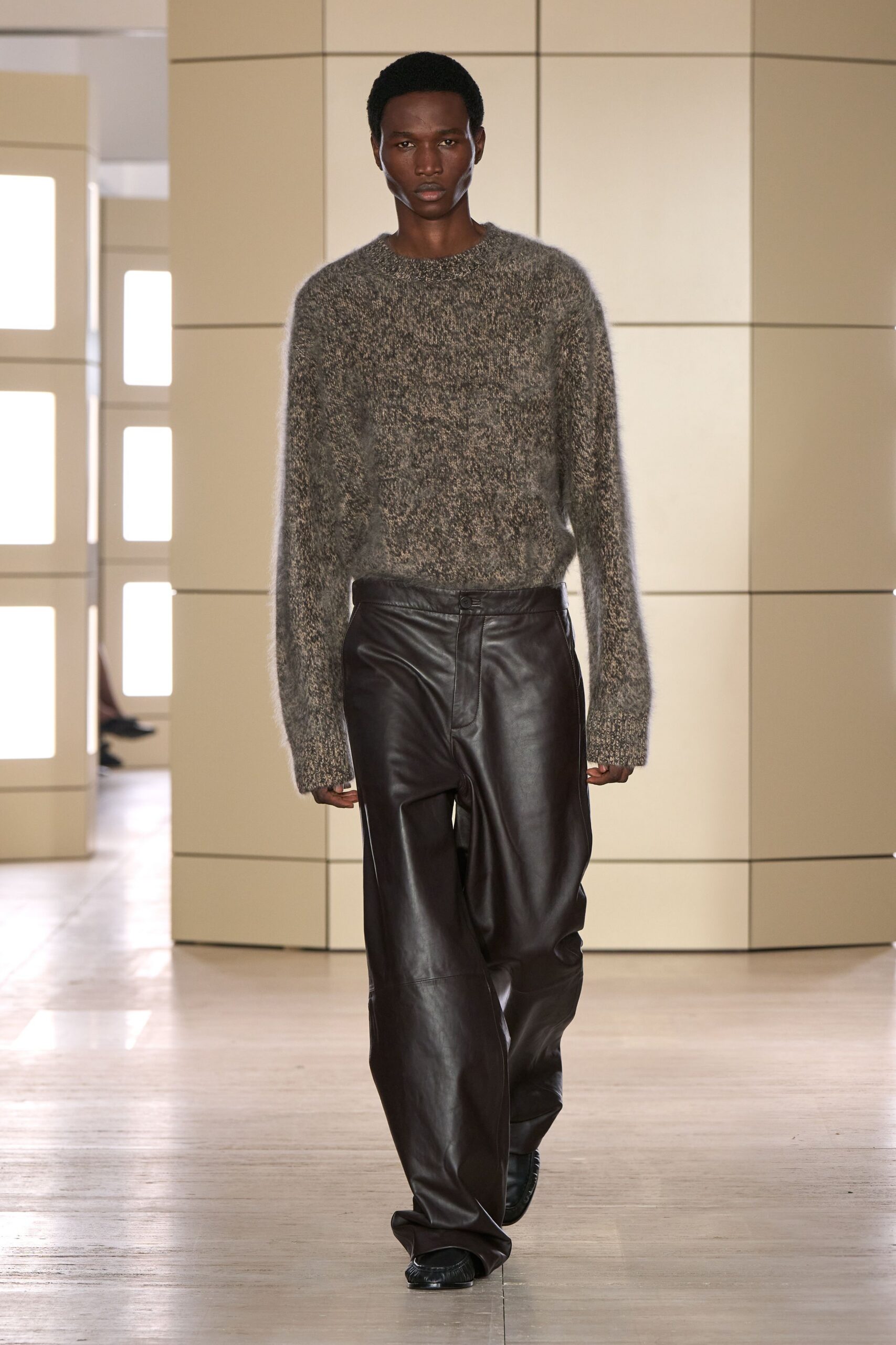 Mottled grey fuzzy knit sweater, sleeves hanging past the knuckles. Dark espresso leather trousers cut wide and pooling over the shoe. Black square-toe shoes peeking from the break. Office atrium at dusk, carrying the heat of the day outside in.