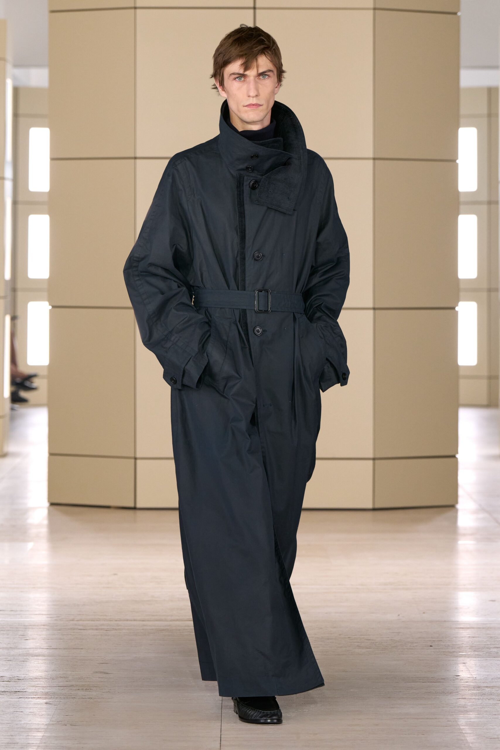 Black knit turtleneck tight to the throat. Ink-dark belted overcoat with a padded stand collar folding across three buttons. Square-toe black leather shoes, lightly creased at the vamp. Crossing a near-empty terminal before dawn with one gate change left.