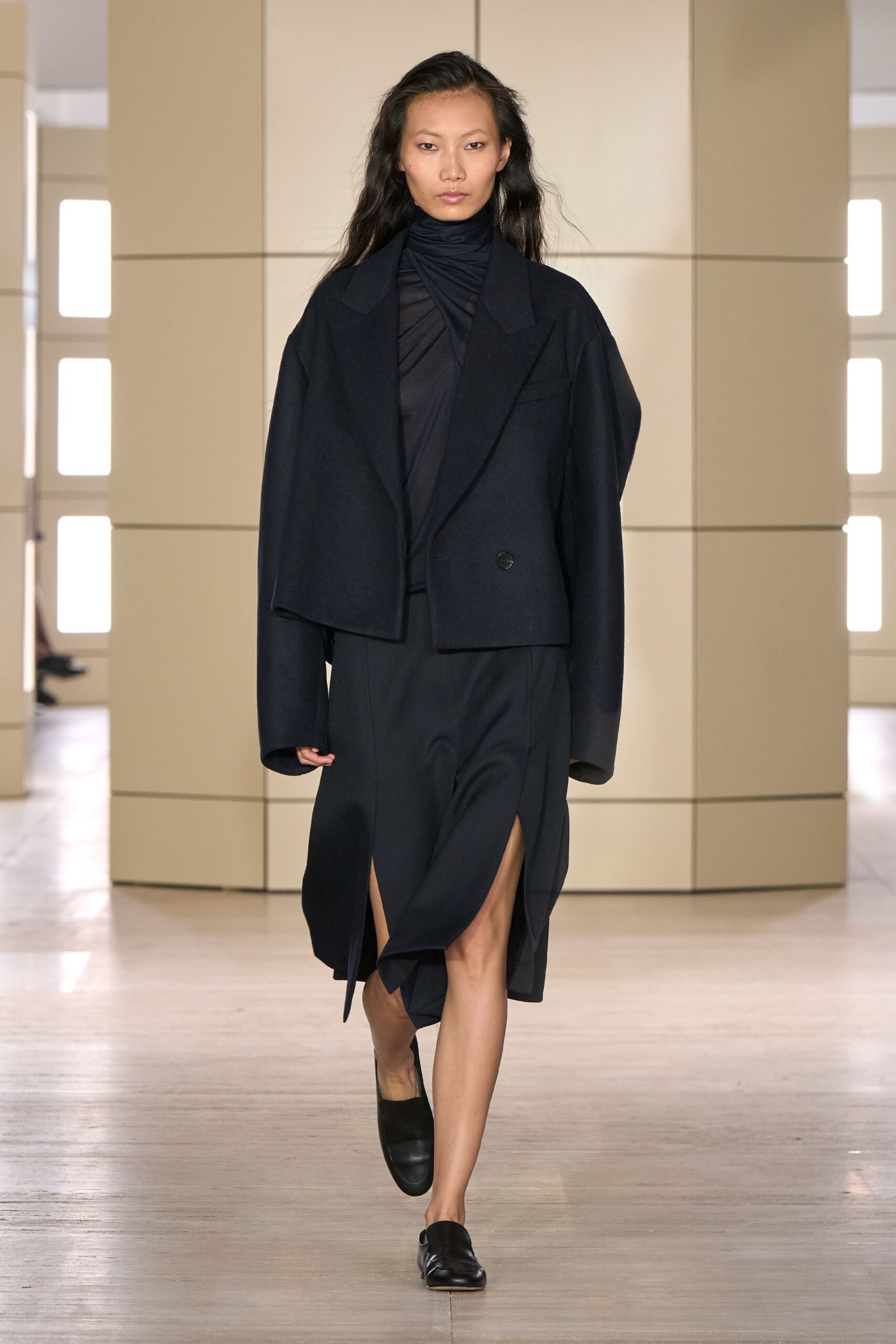 Boxy black wool jacket, one dark button low on the front. Black jersey dress with a ruched funnel neck and wrapped bodice. Asymmetric skirt panel opening to a high slit at one thigh. Black leather loafers with squared, blunt toes. Crossing a silent hotel lobby before the breakfast tables are laid.
